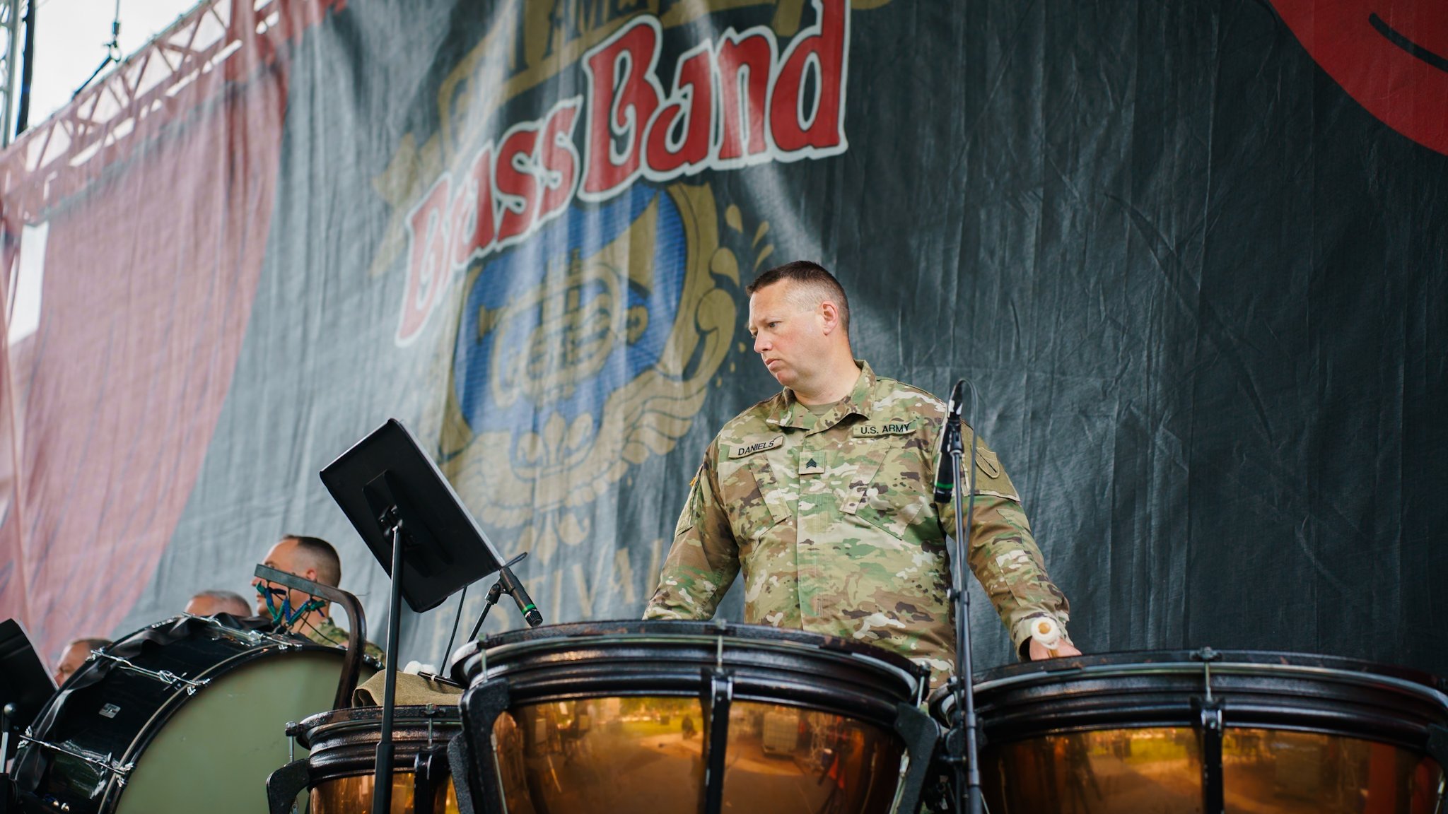 A male soldier in camouflage uniform is playing drums on a stage with a black backdrop featuring a large patriotic logo. Several other drummers are partially visible in the background in Danville, Kentucky.