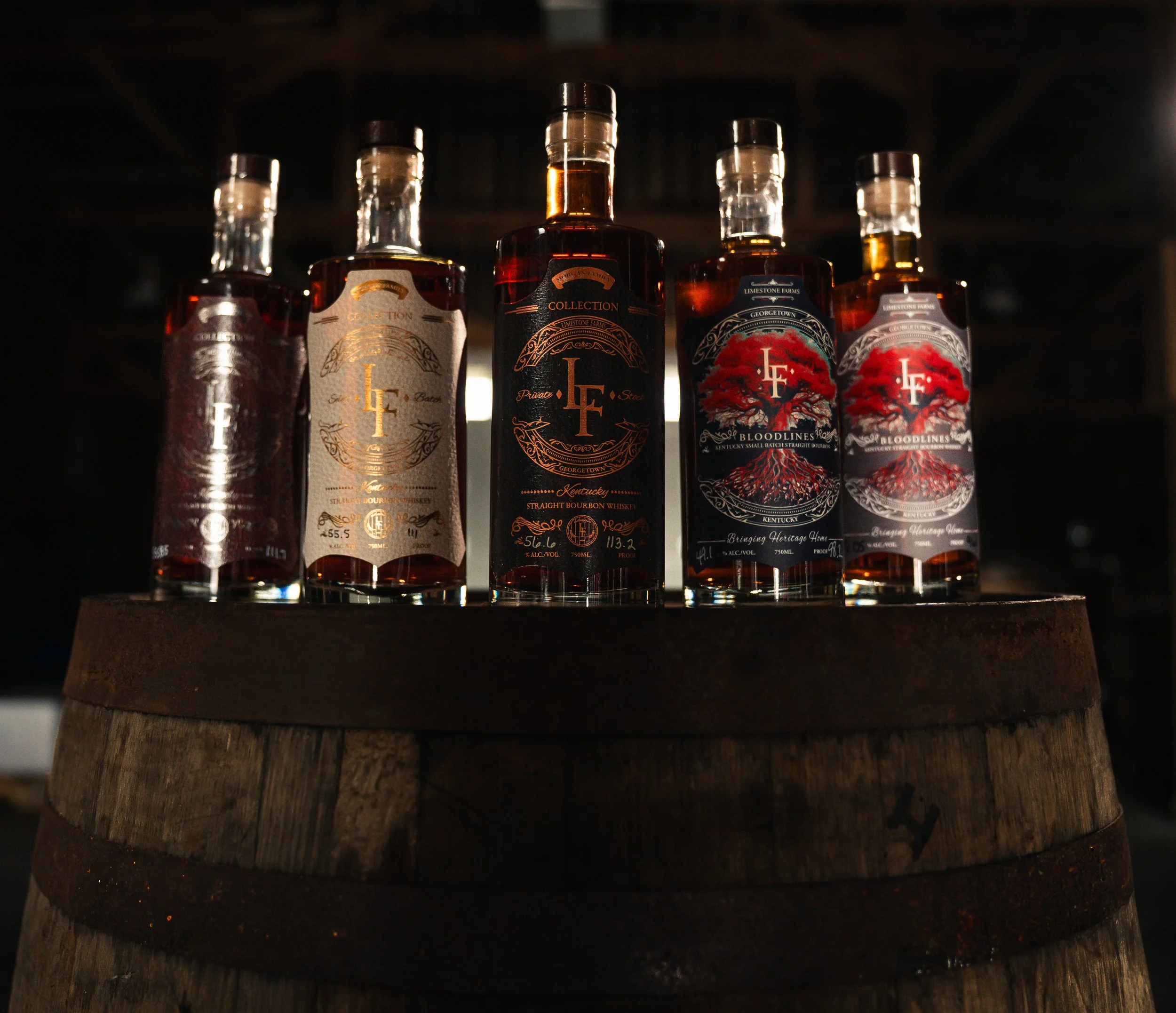 Five bottles of bourbon whiskey placed on top of a wooden barrel in a dimly lit indoor setting.