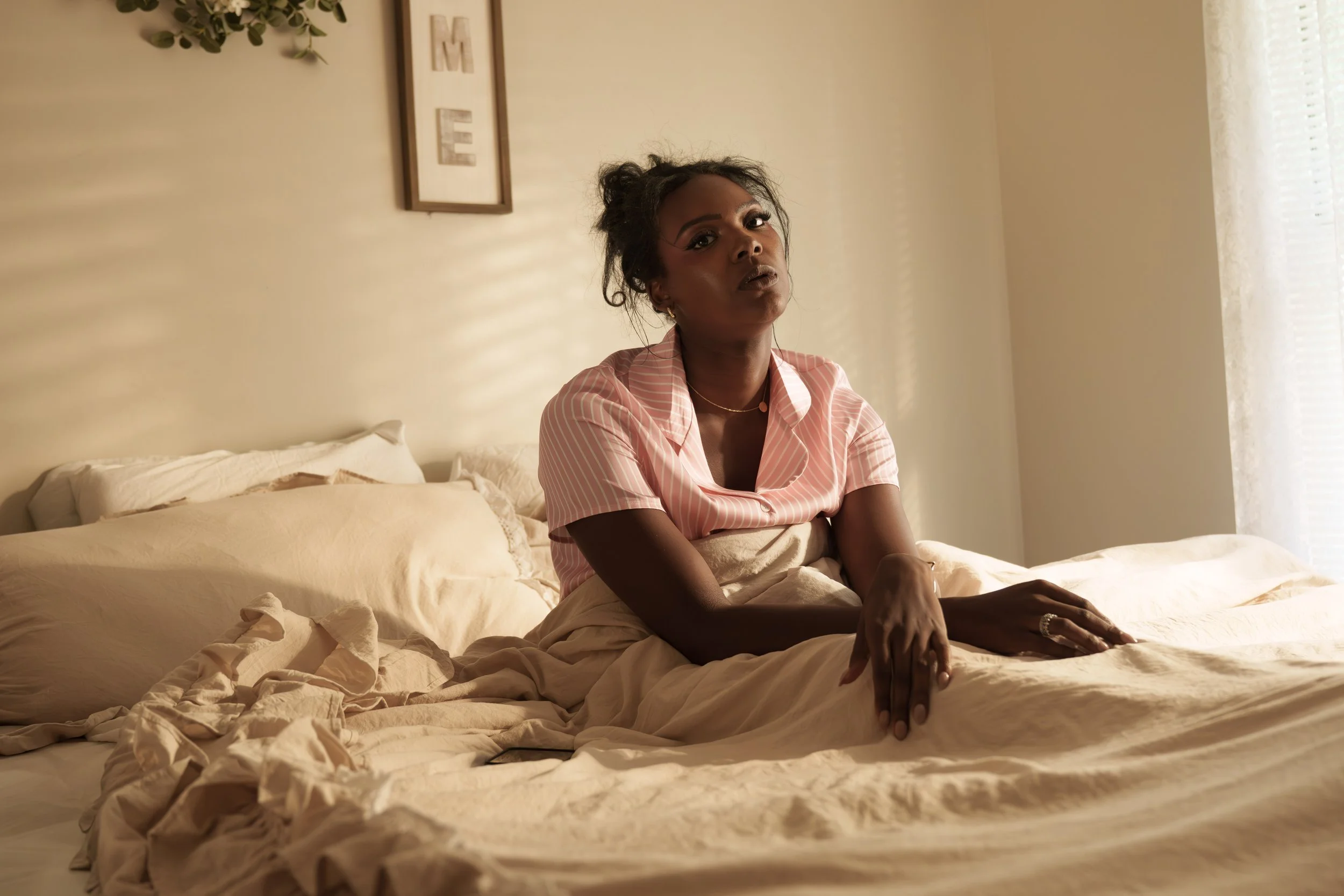 A woman sits on a bed in a softly lit room, wearing pink and white striped pajamas, looking contemplative.
