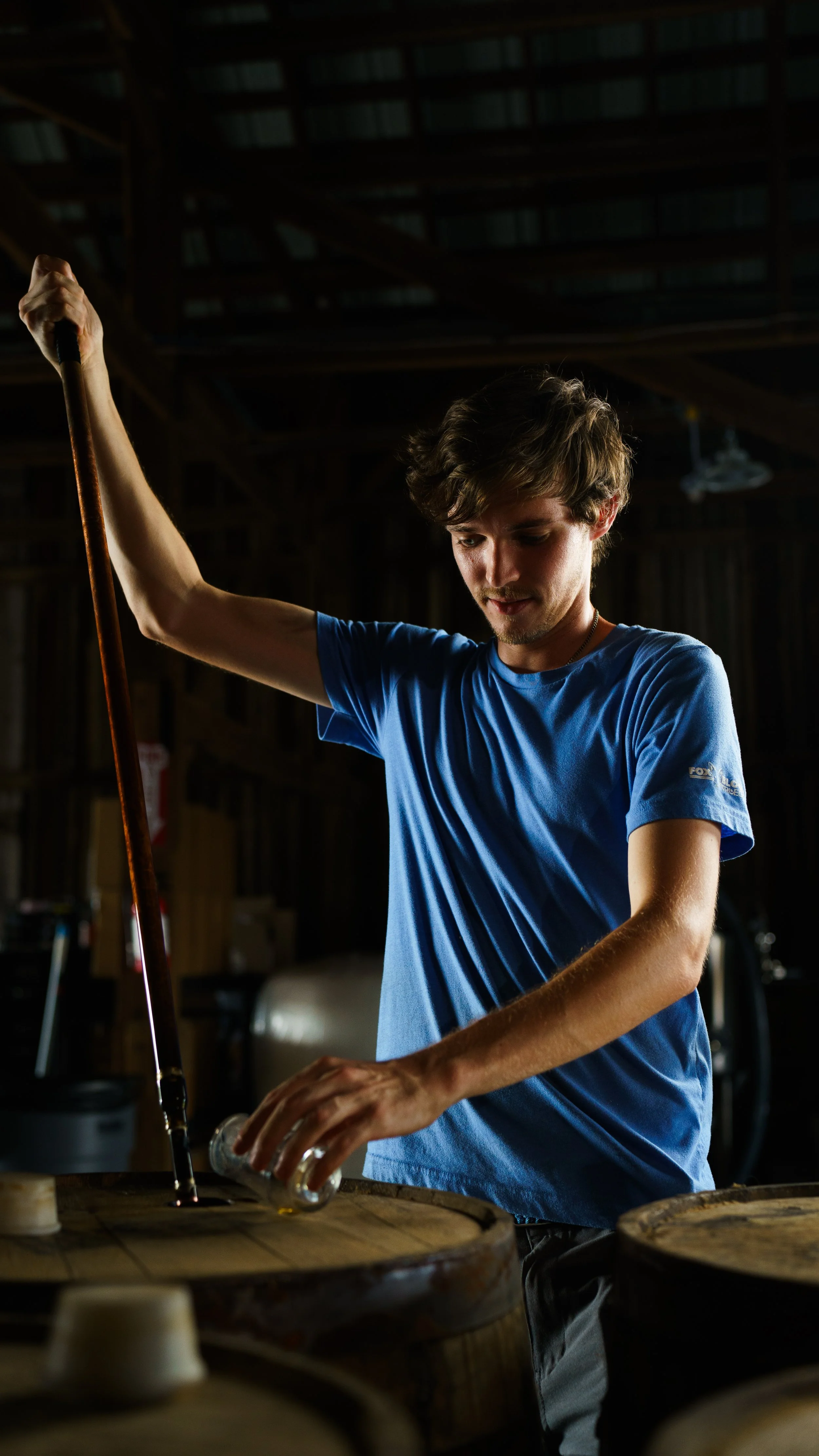 A young man in a blue t-shirt pouring liquid into a wooden barrel inside a dimly lit storage or warehouse space.