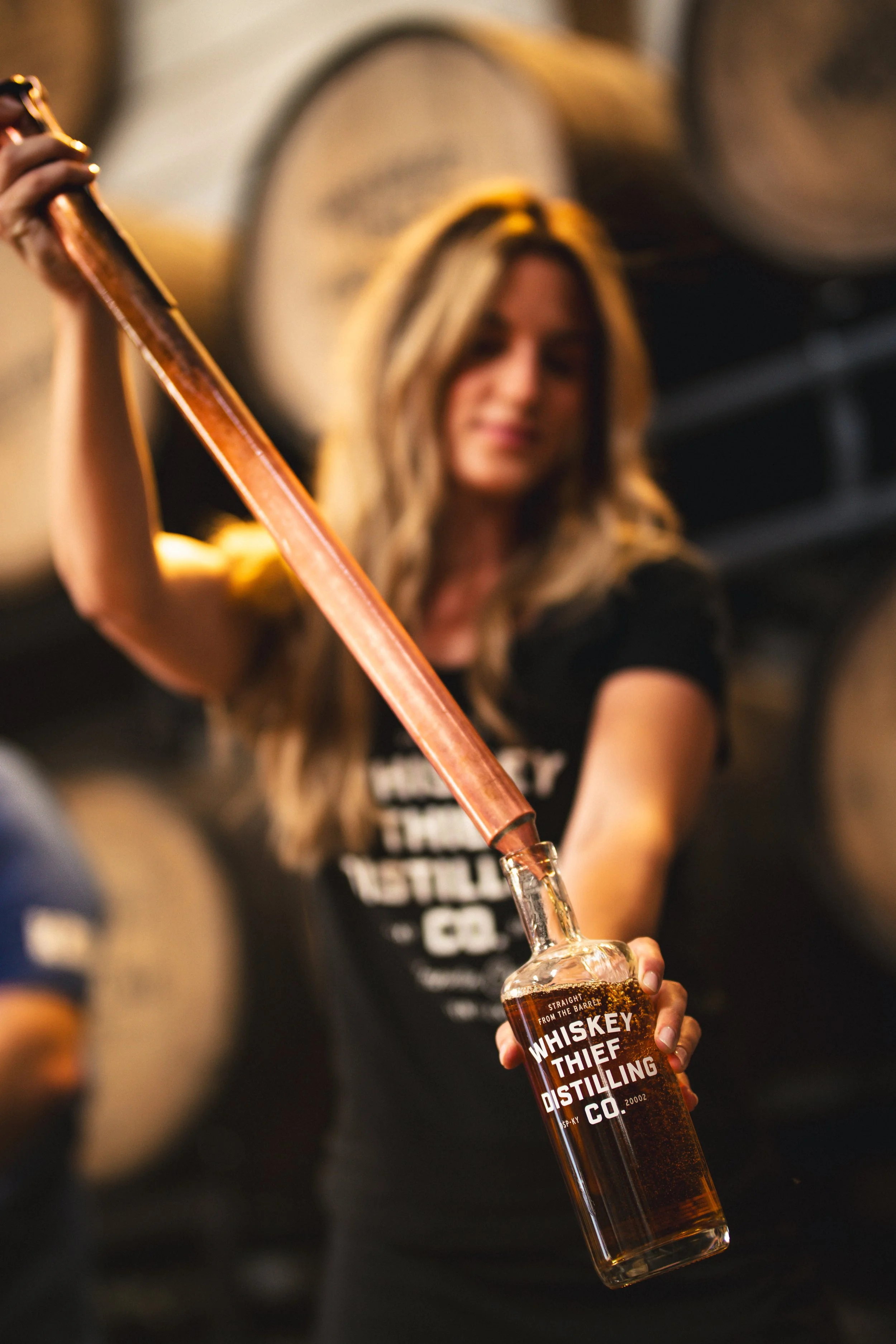 A woman in a black shirt pouring whiskey from a copper still into a bottle labeled 'Whiskey Thief Distilling Co.' against a background of whiskey barrels.