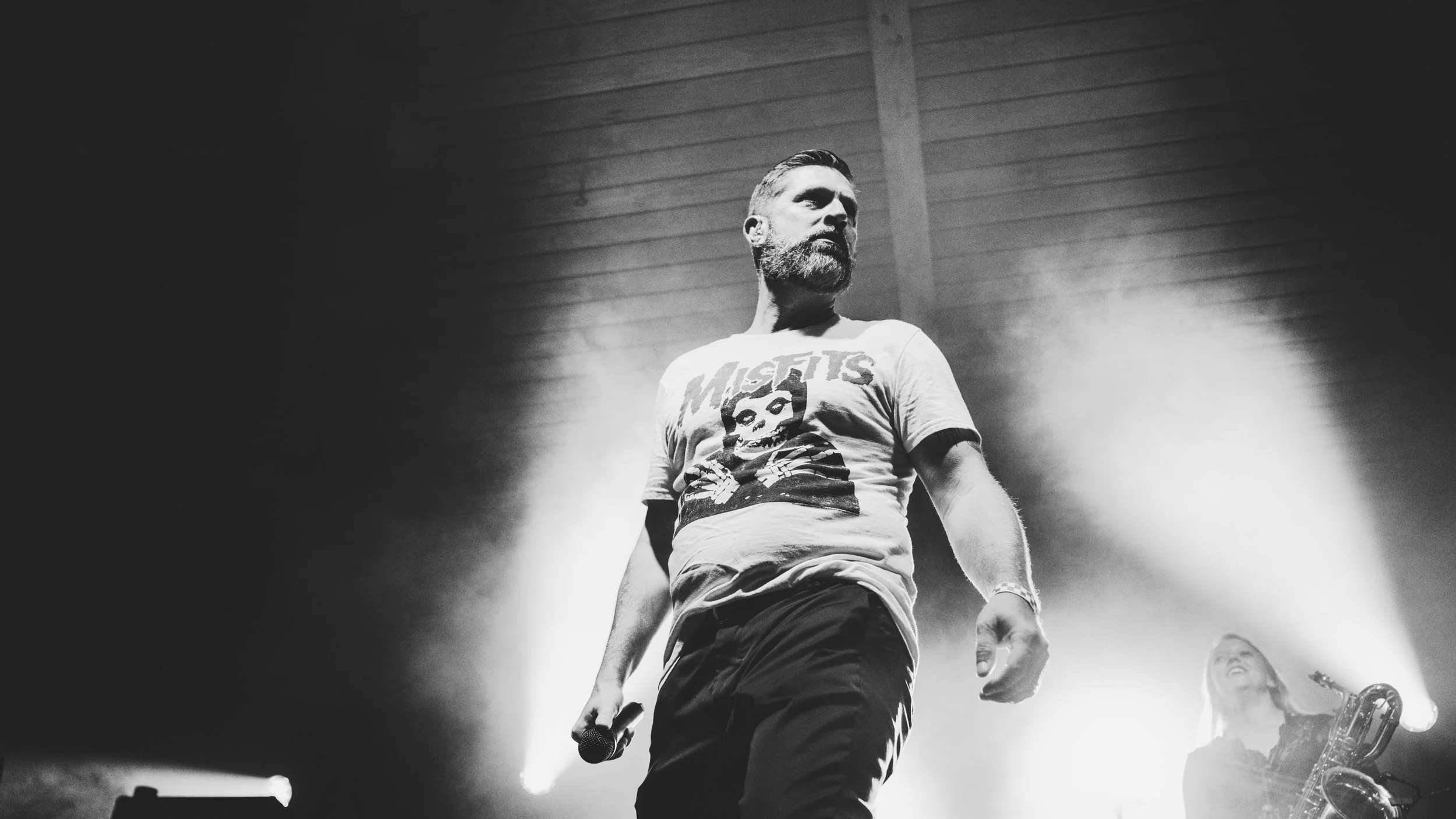 A male performer with a beard and styled hair on stage, holding a microphone in his right hand, wearing a t-shirt with a skull graphic and the text 'Misfits', illuminated by bright stage lights.
