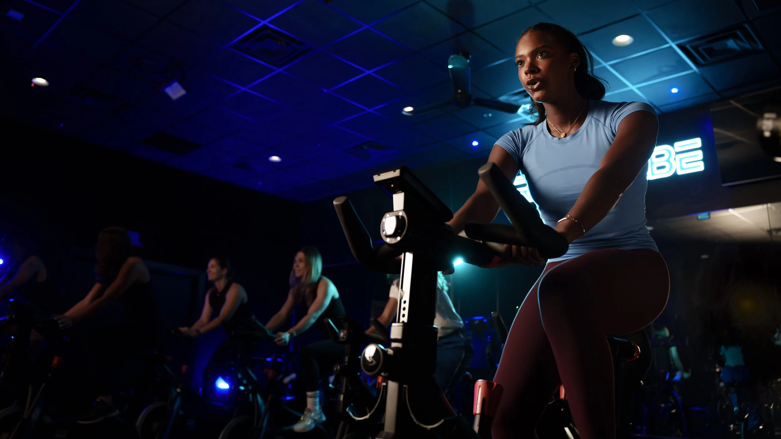 A woman with braided hair is using a stationary exercise bike in a dimly lit indoor gym, surrounded by other women also biking, with blue neon lights and a dark ceiling.