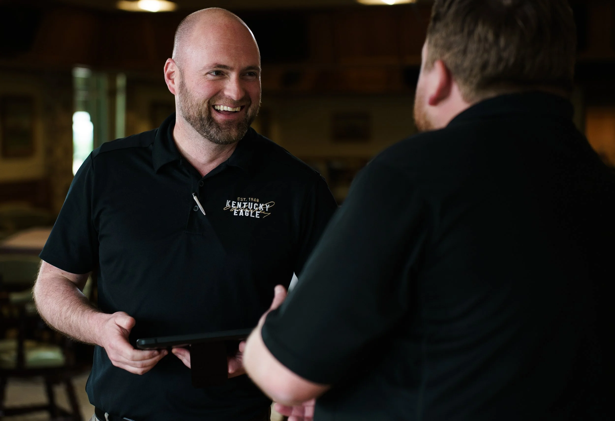 Two men in black shirts, smiling and talking indoors. The man on the left has a shaved head and a beard, holding a tablet. The other man has short hair and is facing away. The shirts have a Kentucky Eagle logo.