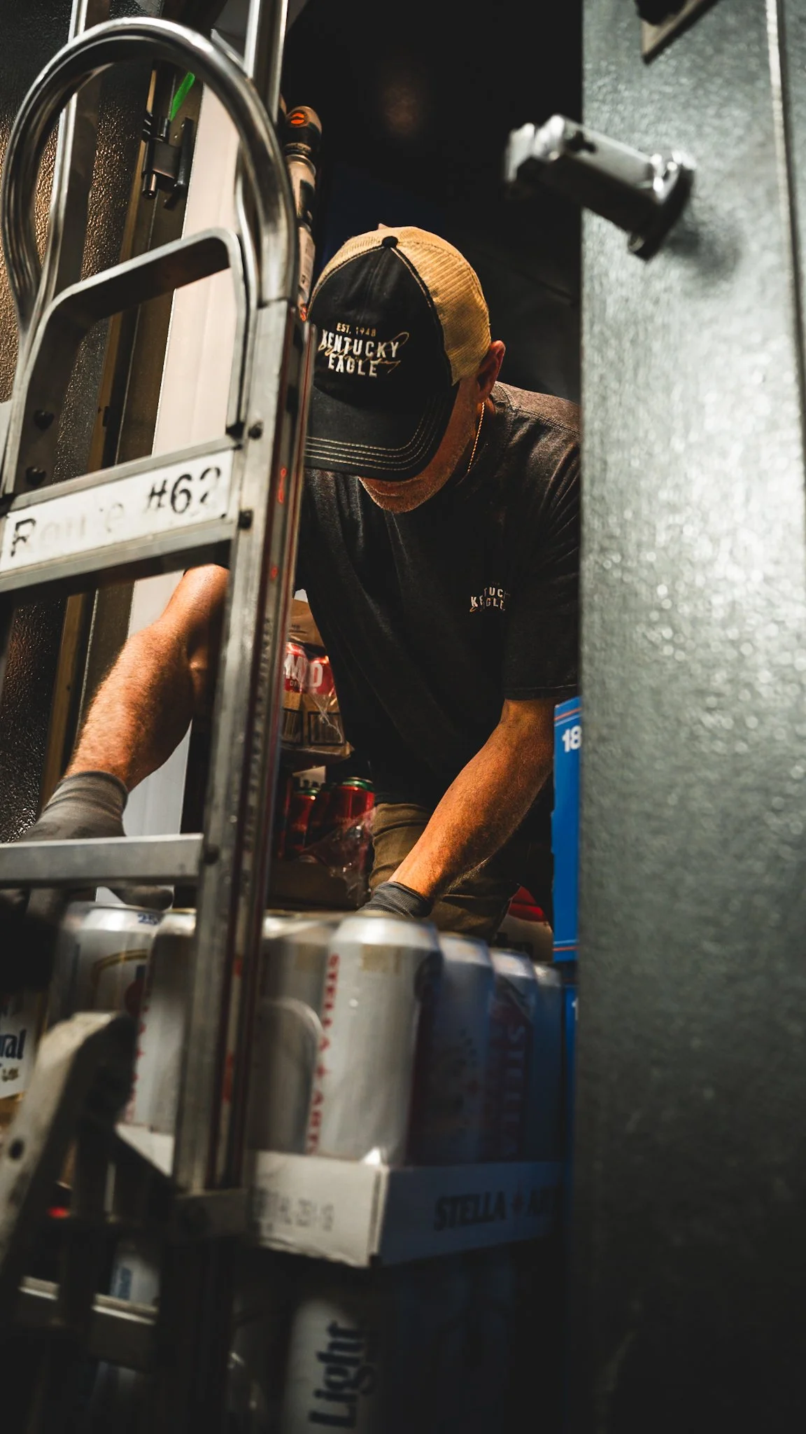A man wearing a black and tan Kentucky Eagle baseball cap and a black T-shirt is working in a storage room, organizing cans of drinks on shelves from a low angle view.