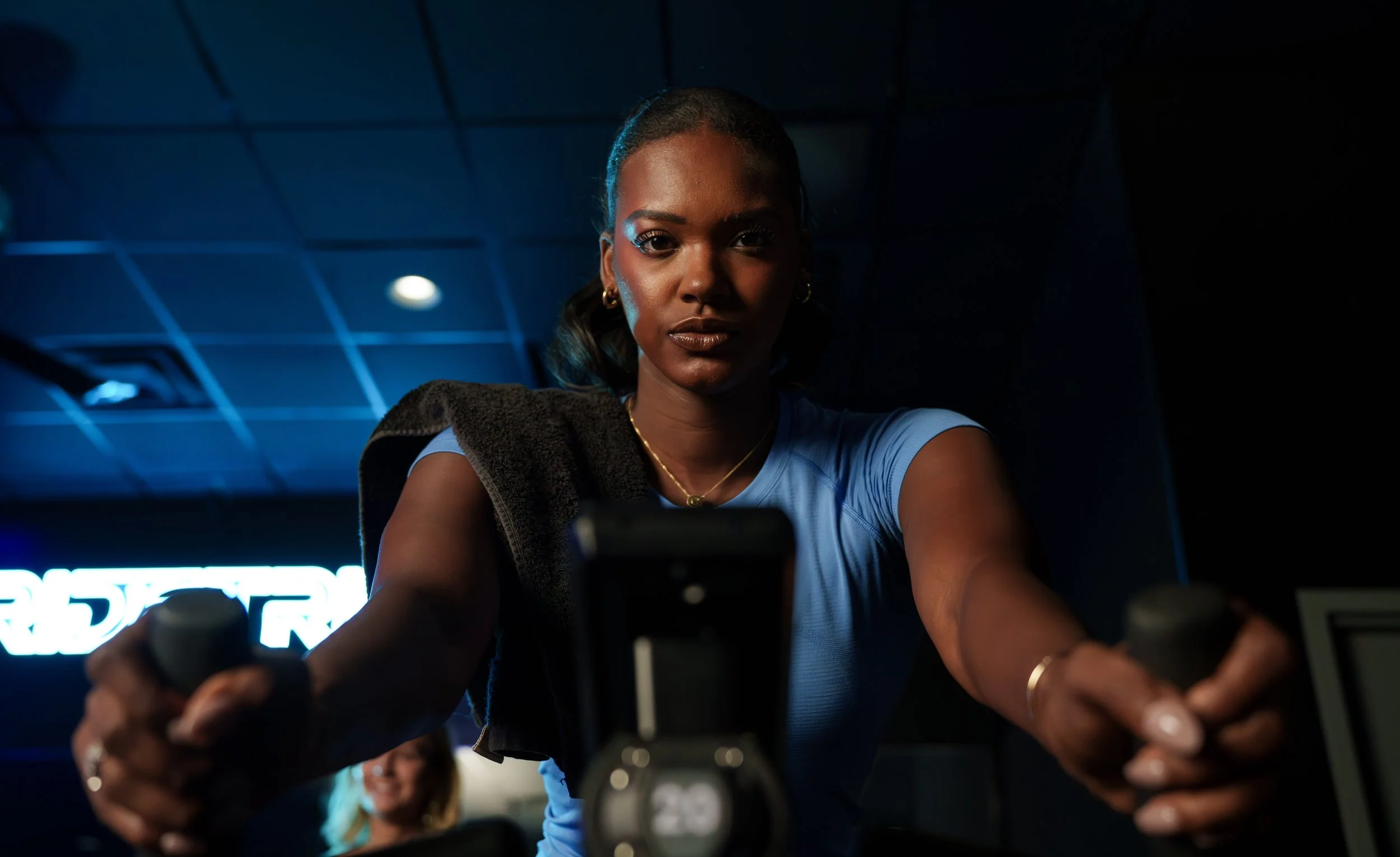 A woman in a blue shirt is focused as she exercises on a bike, with a towel draped over her shoulder, in a dimly lit fitness center. She is looking at her phone mounted on the bike, with neon lights in the background.