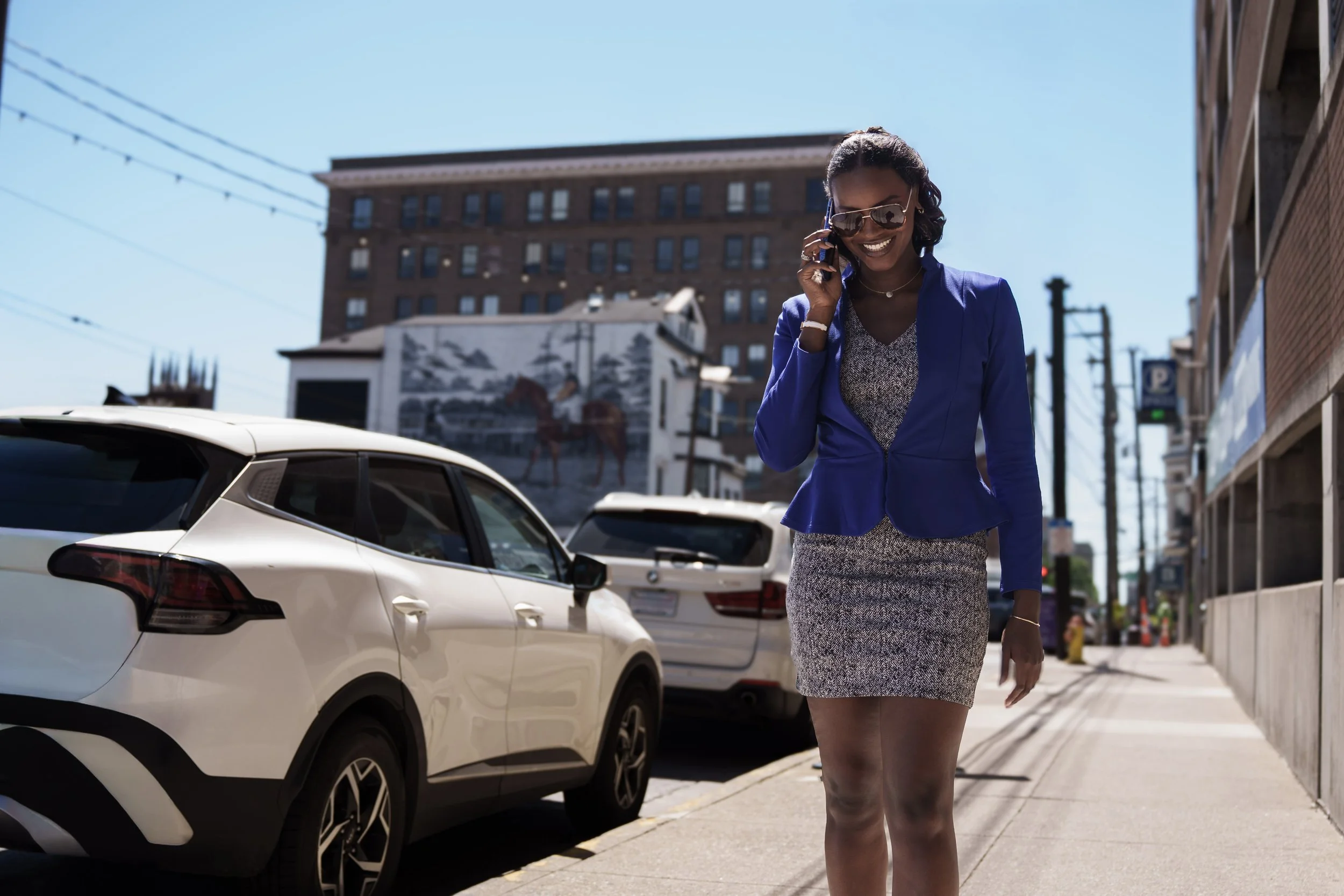 A woman wearing a blue blazer and patterned dress walking on city sidewalk while talking on her cell phone, with parked white cars and tall buildings in the background on a sunny day.