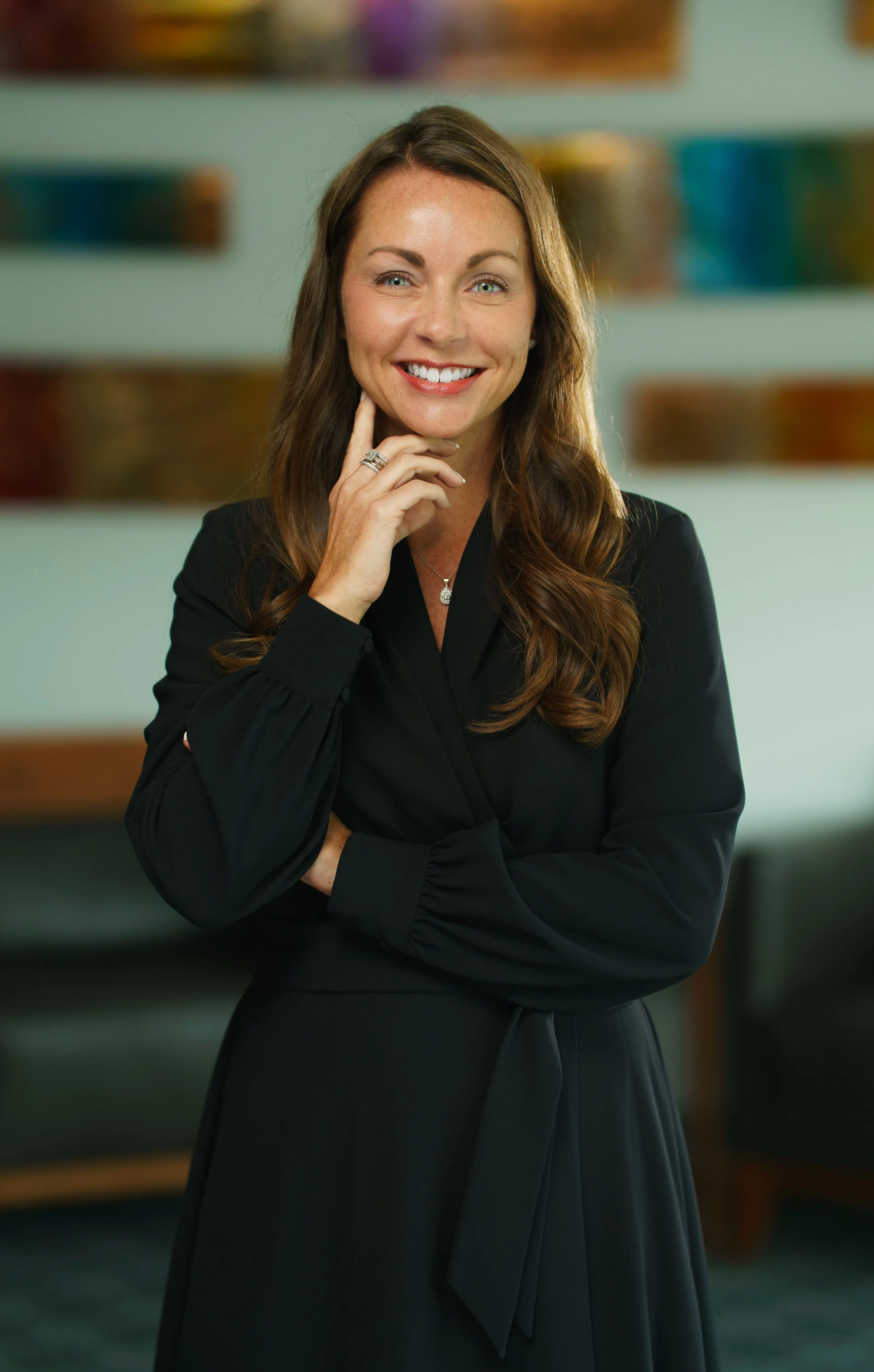 A woman with long brown hair wearing a black dress, smiling, standing with her hand on her chin.