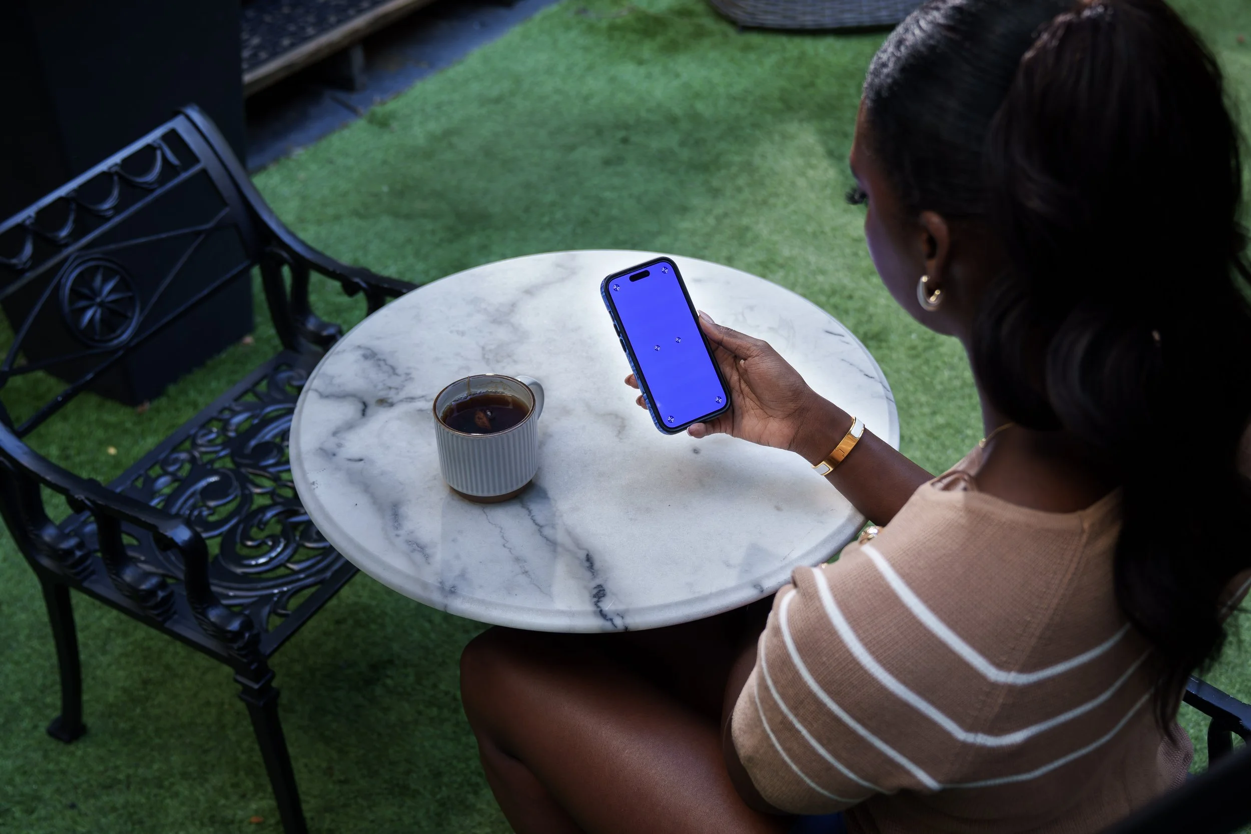 A woman sitting at a small round marble table outdoors, holding a smartphone with a blue screen, with a cup of coffee on the table, on a grassy area with black wrought iron chairs around.