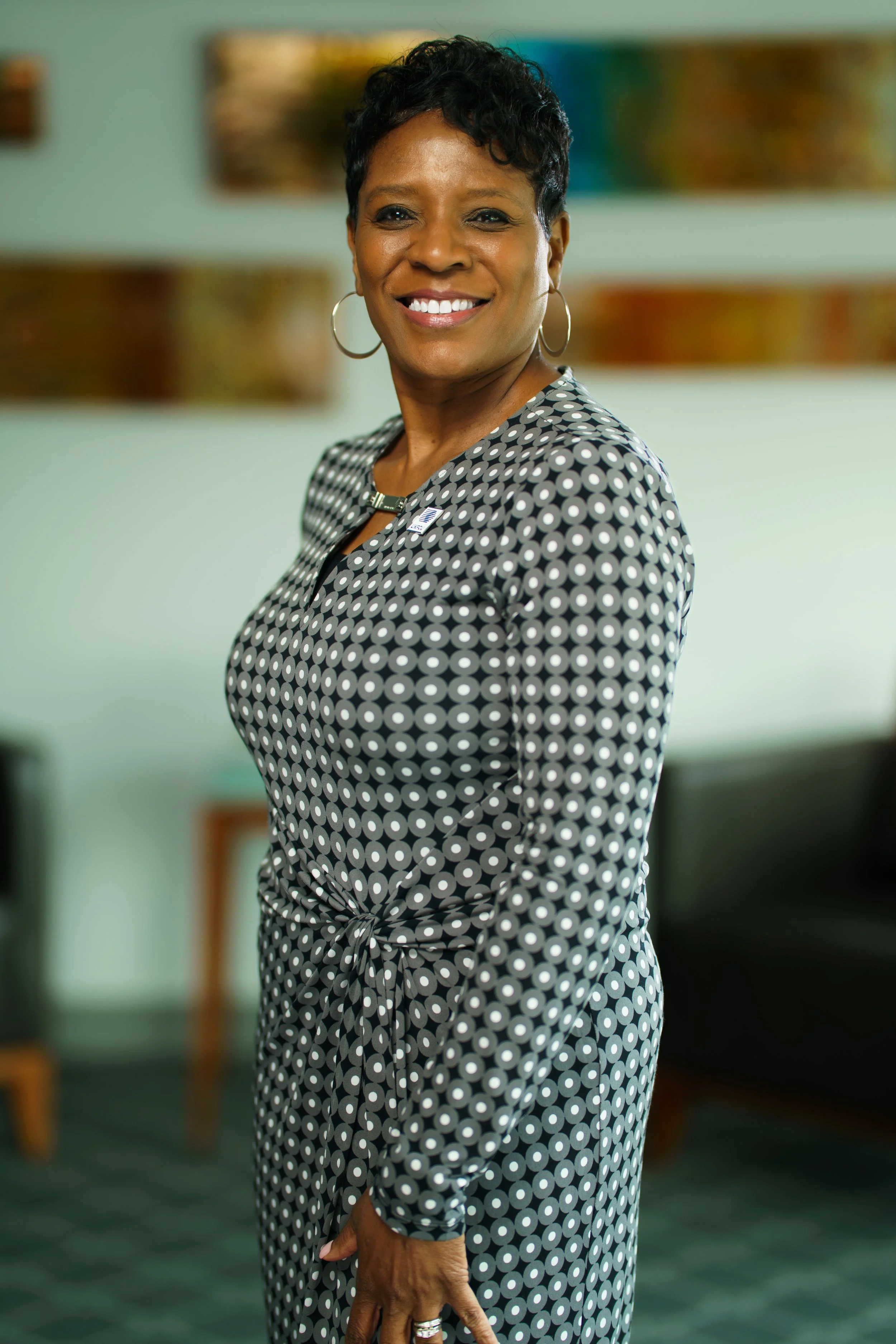 A smiling African American woman with short curly hair wears a black and white polka dot dress with earrings, standing in a room with colorful artwork in the background.