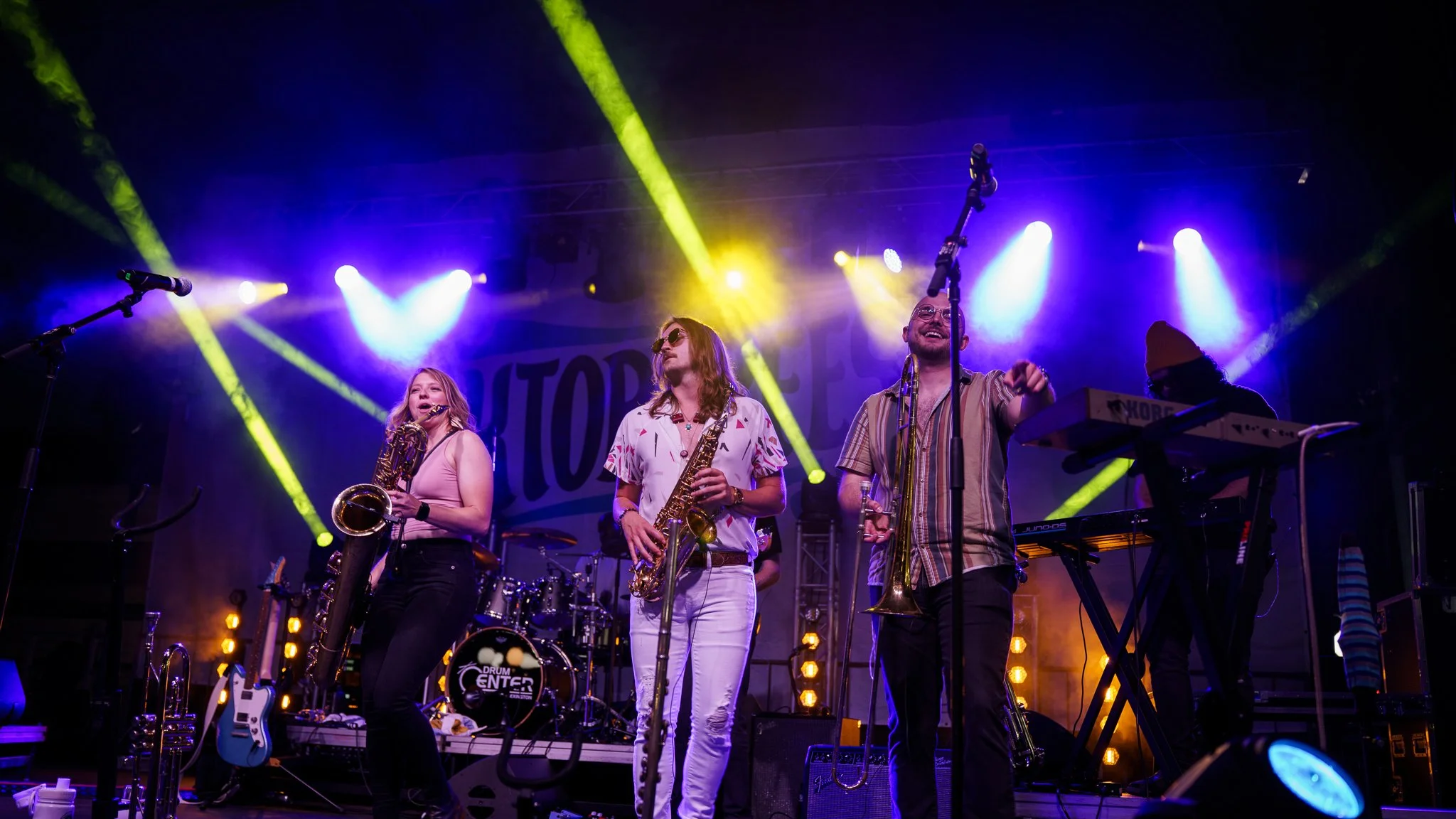 A band performing live on stage with three musicians playing wind instruments and one on keyboard, with colorful stage lighting.