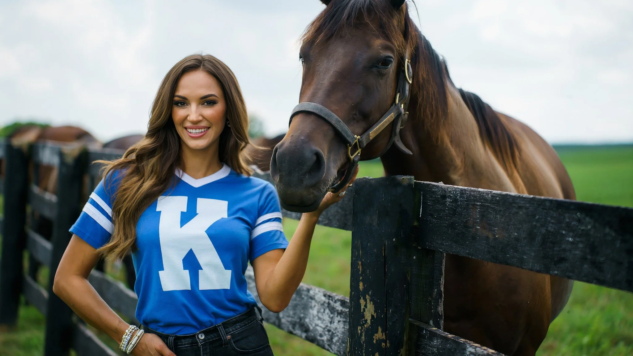 A woman with long brown hair wearing a blue sports jersey with a large white letter 'K' on it, smiling and petting a brown horse near a wooden fence in a grassy field.
