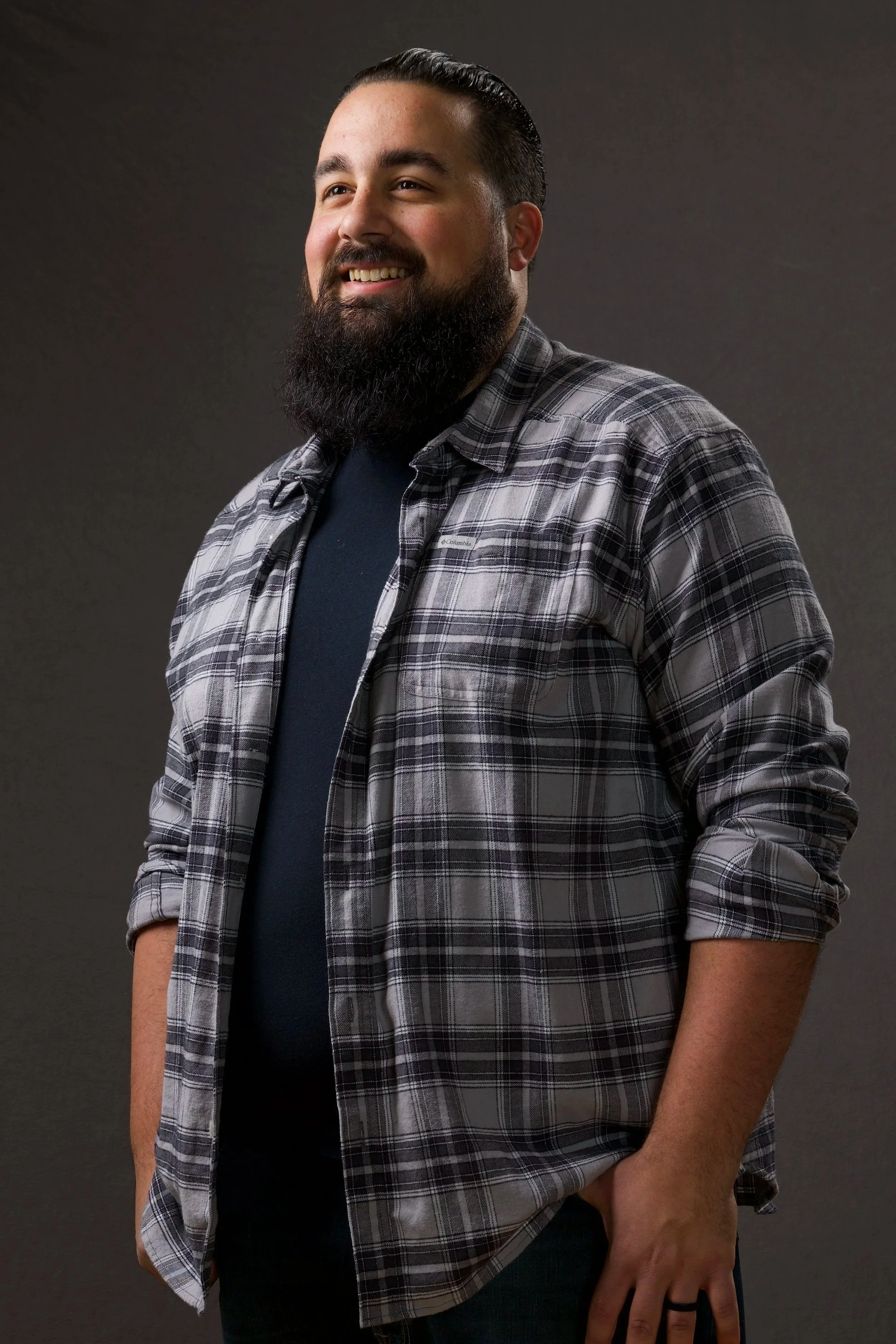A man with a beard and mustache, smiling and looking slightly to the side. He is wearing a black T-shirt and a gray plaid shirt over it, with the sleeves rolled up. His left hand is in his pocket, and he is standing against a plain, dark background.