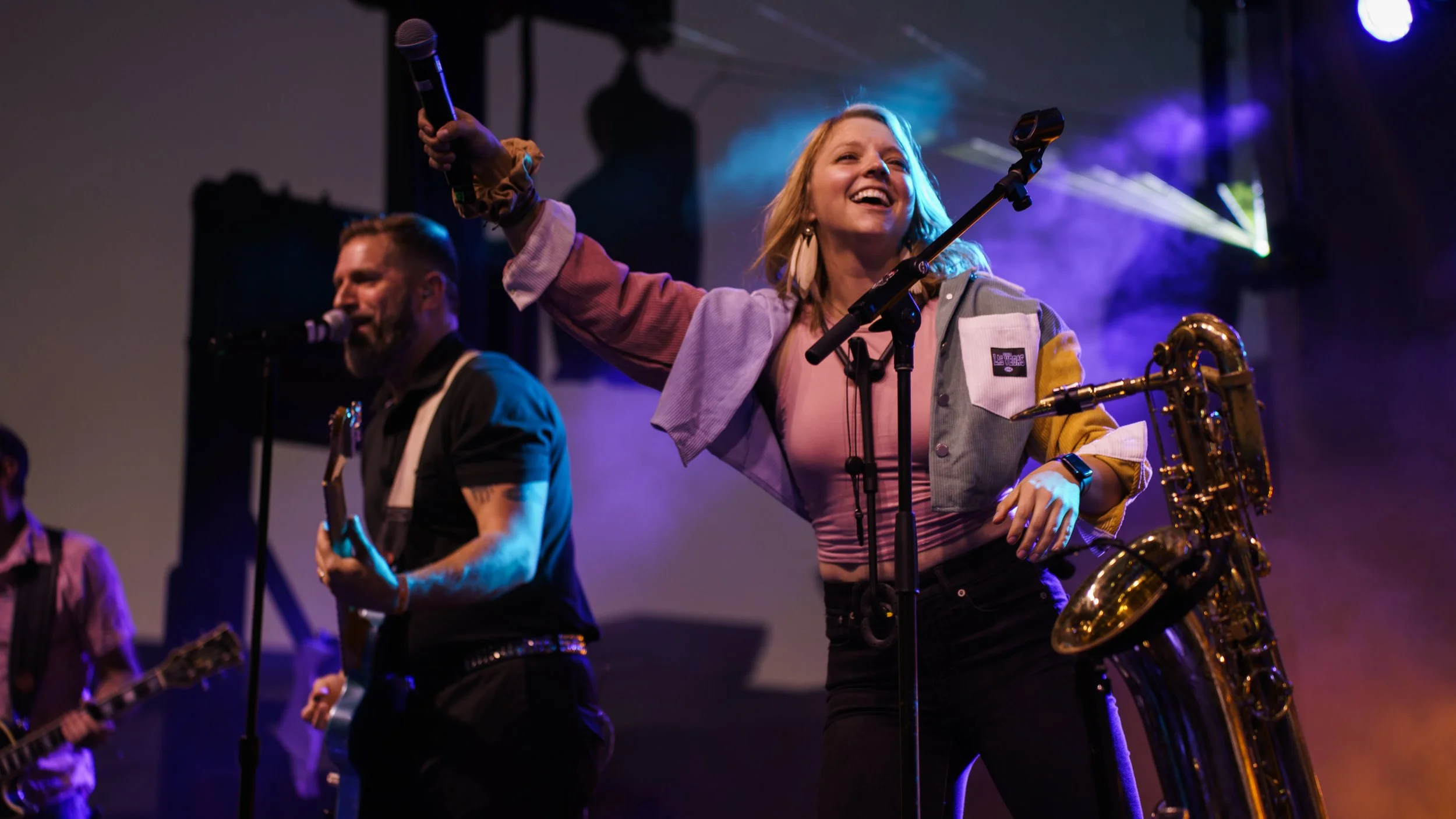 A group of musicians performing on stage, with a woman singing into a microphone and holding a saxophone, a man playing guitar in the background, and colorful stage lighting.