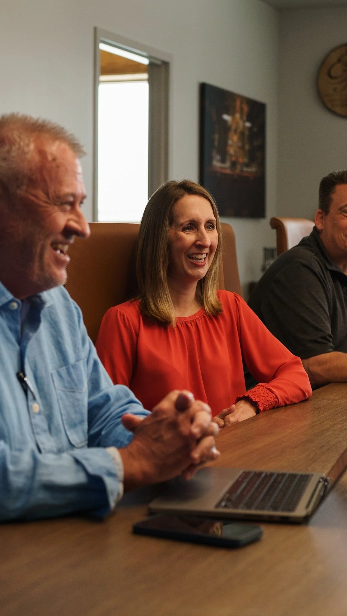 Three adults sitting at a wooden conference table, laughing and smiling, with a laptop and a smartphone in front of them in a meeting room.