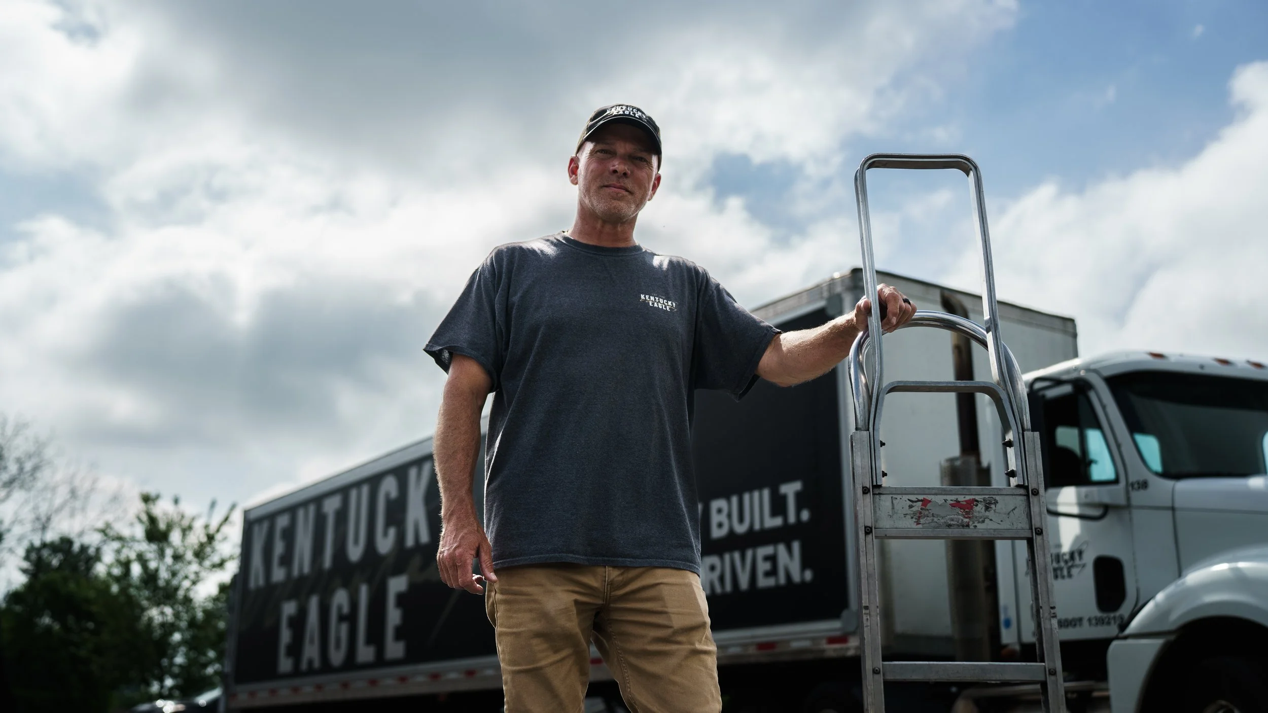 Man standing outdoors near a moving truck with a ladder, wearing a Kentucky Eagle T-shirt, under a cloudy sky.