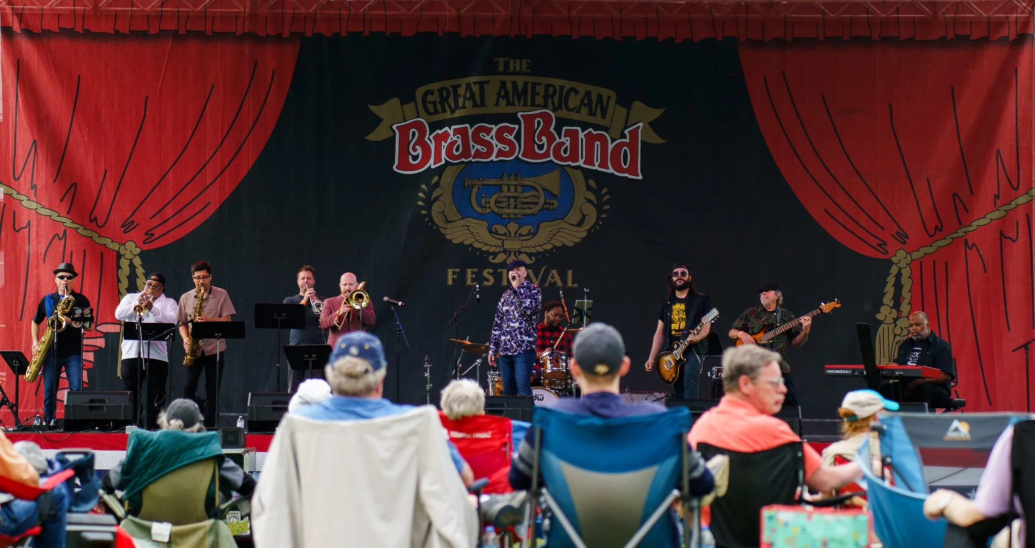 A band performs on stage at the Great American Brass Band Festival with an audience seated in chairs and blankets outdoors.