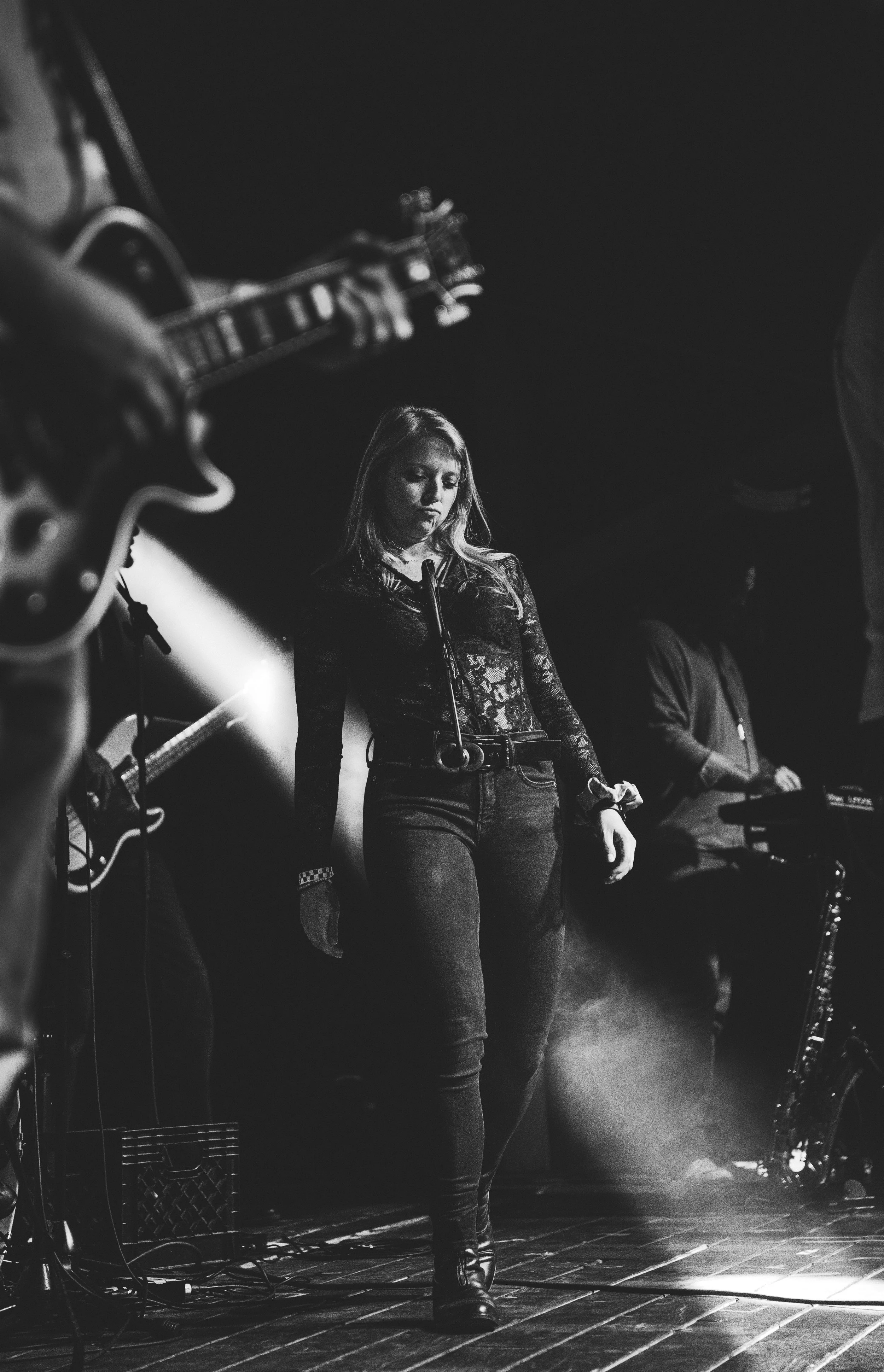 A woman singing on stage with musicians playing guitars and keyboards in the background, in a black and white photo.
