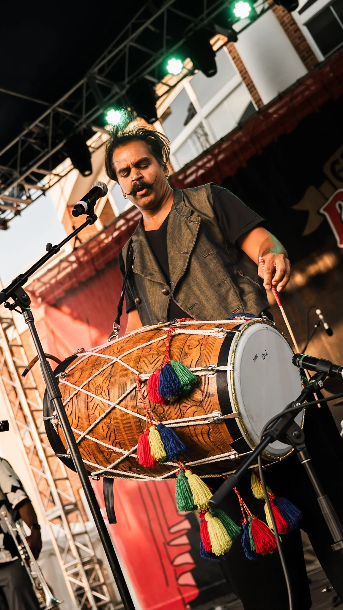A musician with a mustache and dark hair is playing a dhol drum during a live outdoor concert. He is wearing a dark vest and black t-shirt and appears to be focused on his performance. The stage has lighting and a backdrop with red and black colors.