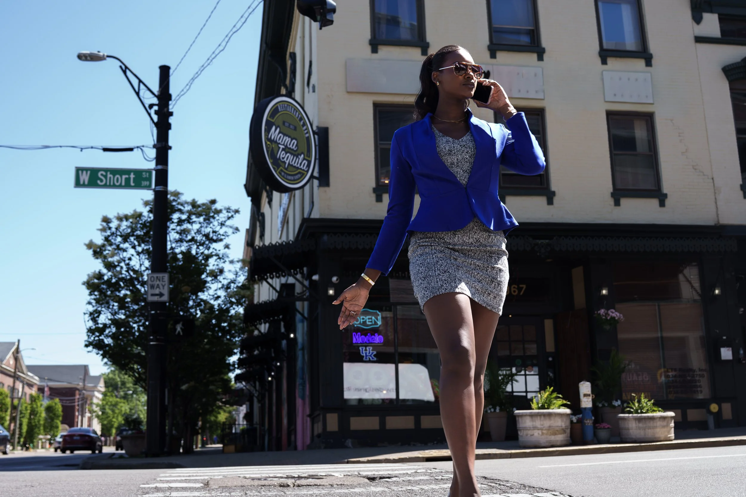 A woman in a blue blazer and patterned skirt walking down the street while talking on her cell phone, with buildings and street signs in the background.