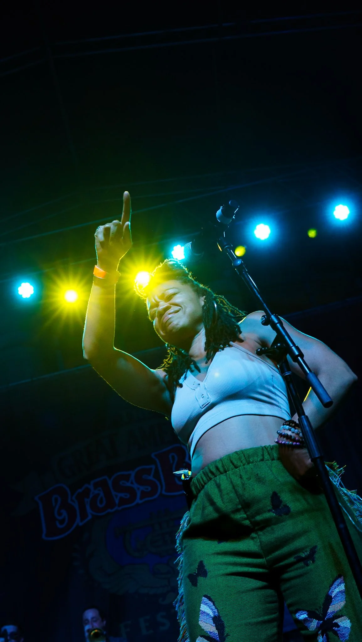 A woman with dreadlocks performing on stage, raising her right hand with her middle finger extended, illuminated by colorful stage lights.