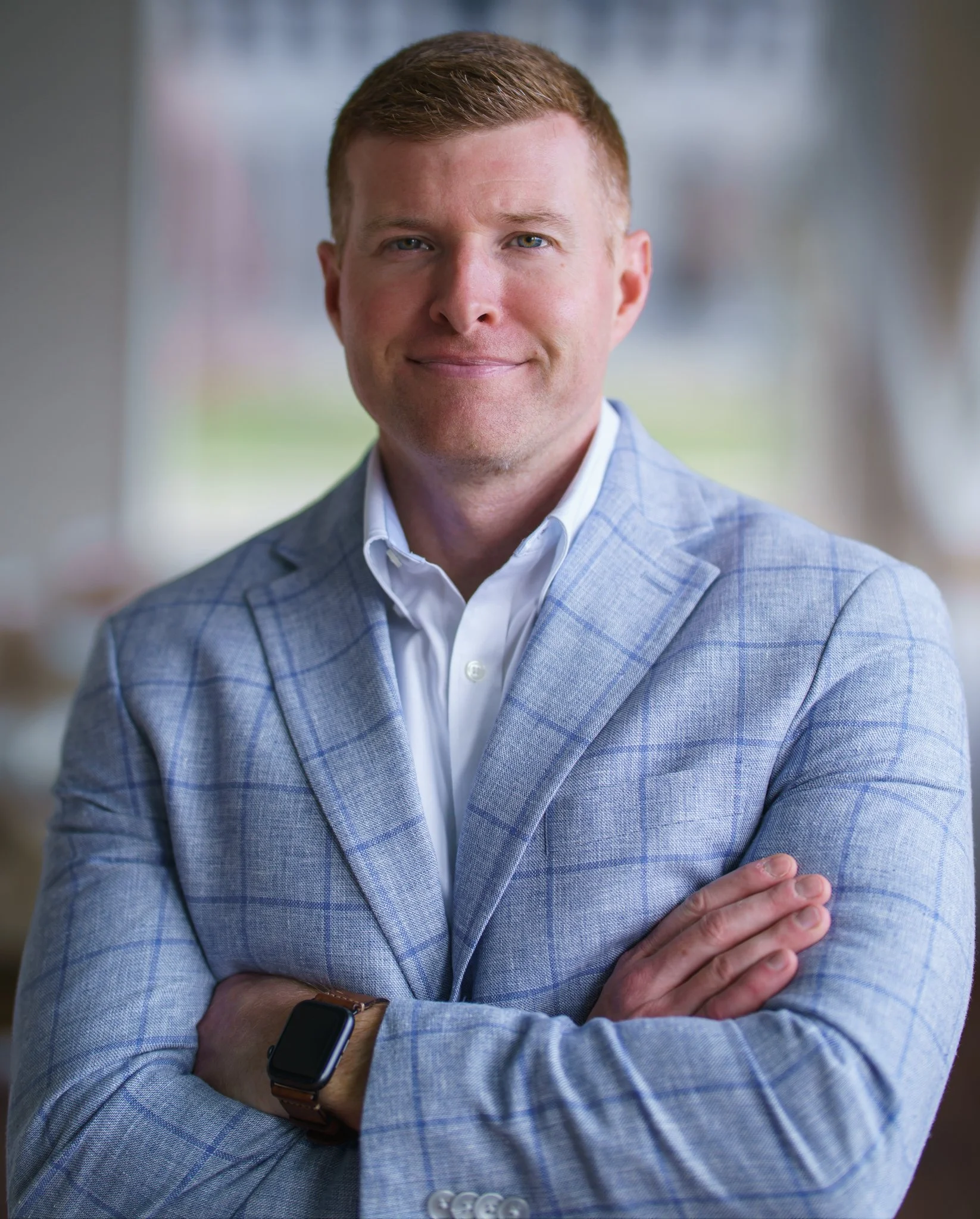 A confident man with crossed arms, wearing a light gray checked blazer, white shirt, and smartwatch, standing indoors with a blurred background.