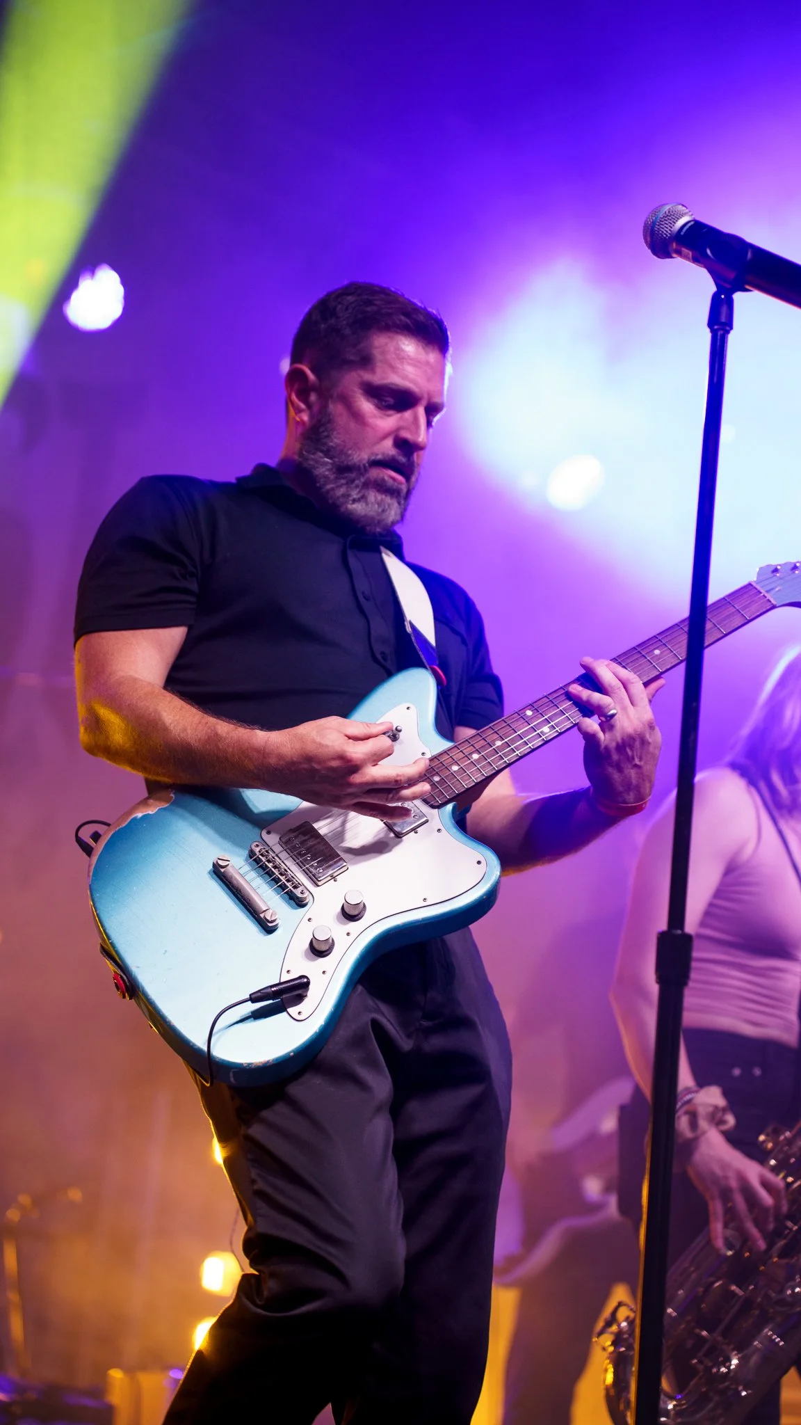 A male guitarist playing an electric guitar on stage with colorful lights and fog in the background.