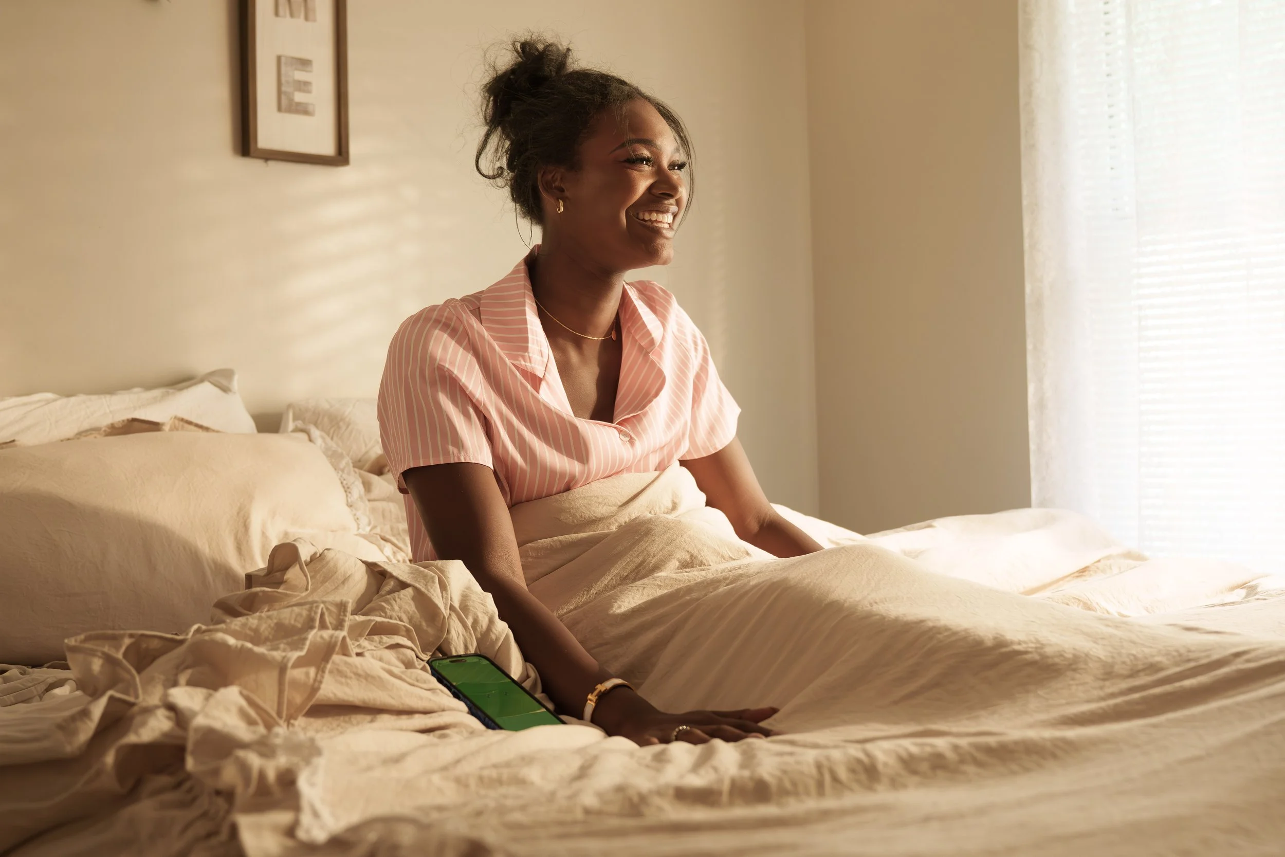 A young woman with dark skin, wearing a pink striped pajama top, is smiling and sitting on a bed near a window with blinds. She has gold hoop earrings and a necklace, and is sitting next to a crumpled comforter and a smartphone with a green screen.