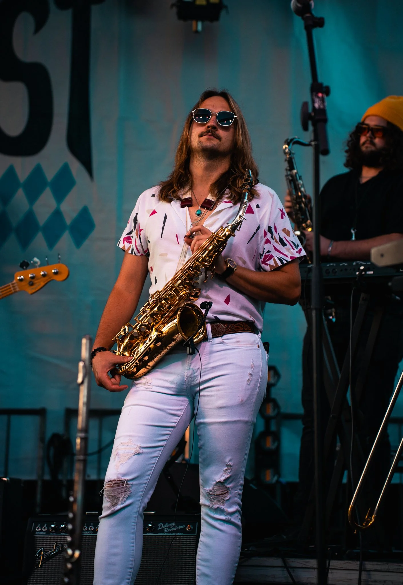 Musician with long hair, sunglasses, and a patterned shirt playing a gold saxophone on stage, with band members and musical equipment in the background.