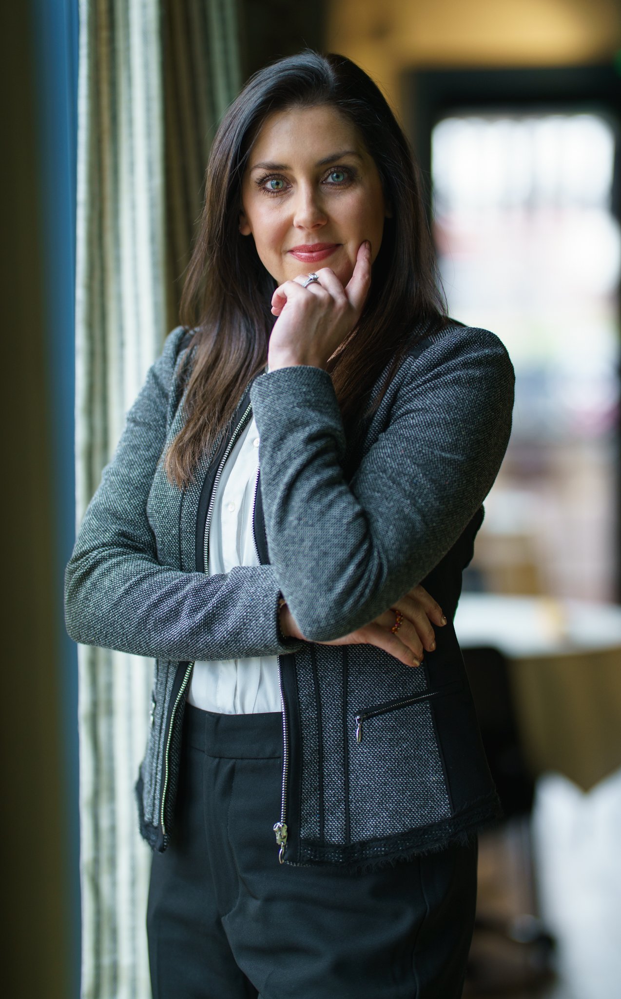 A woman with long dark hair and bright blue eyes standing indoors near a window, wearing a gray blazer and white blouse, with her hand resting on her chin.