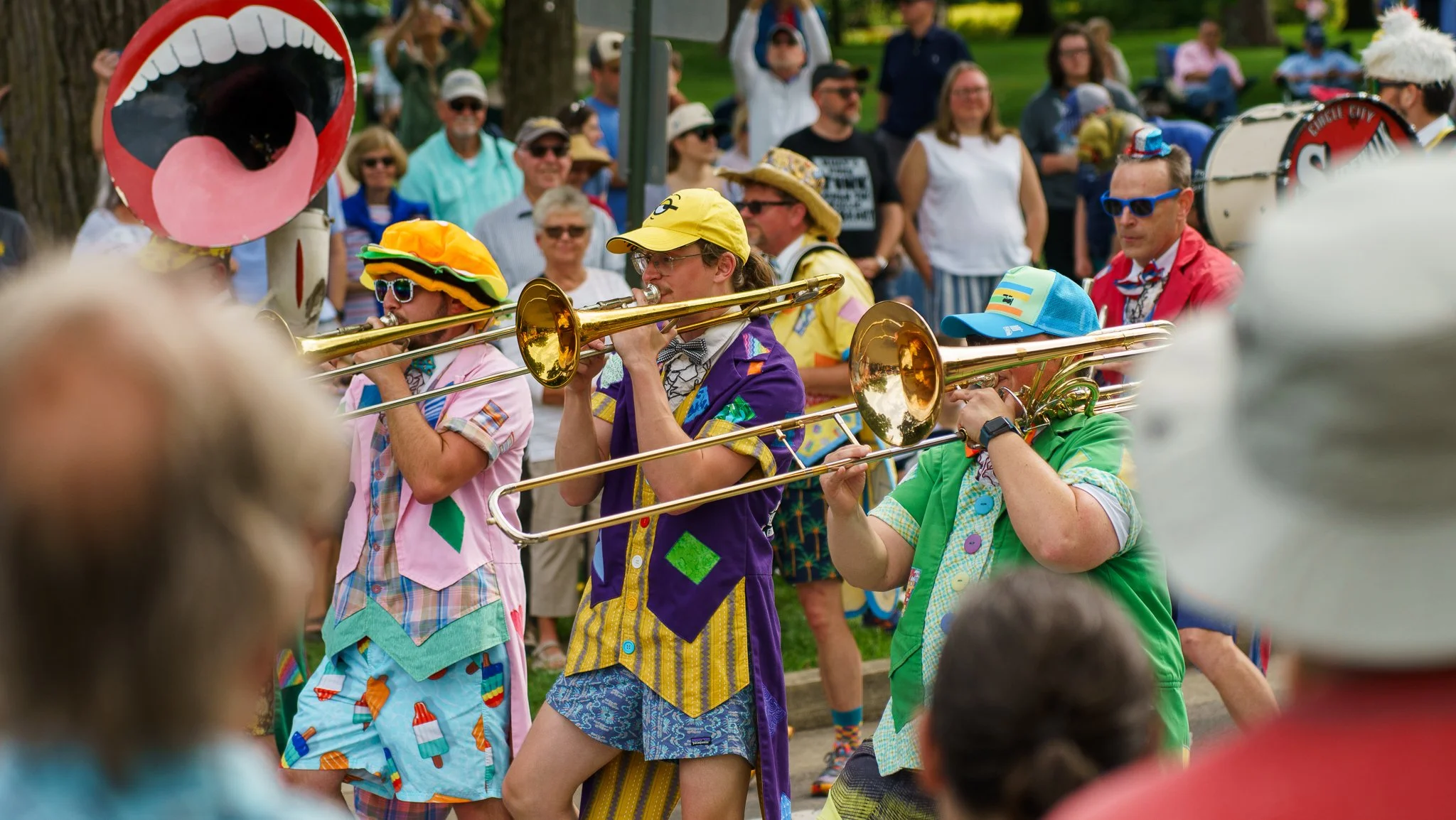 A lively parade scene featuring musicians playing trombones dressed in colorful, playful, and eccentric costumes, with a crowd of spectators watching.