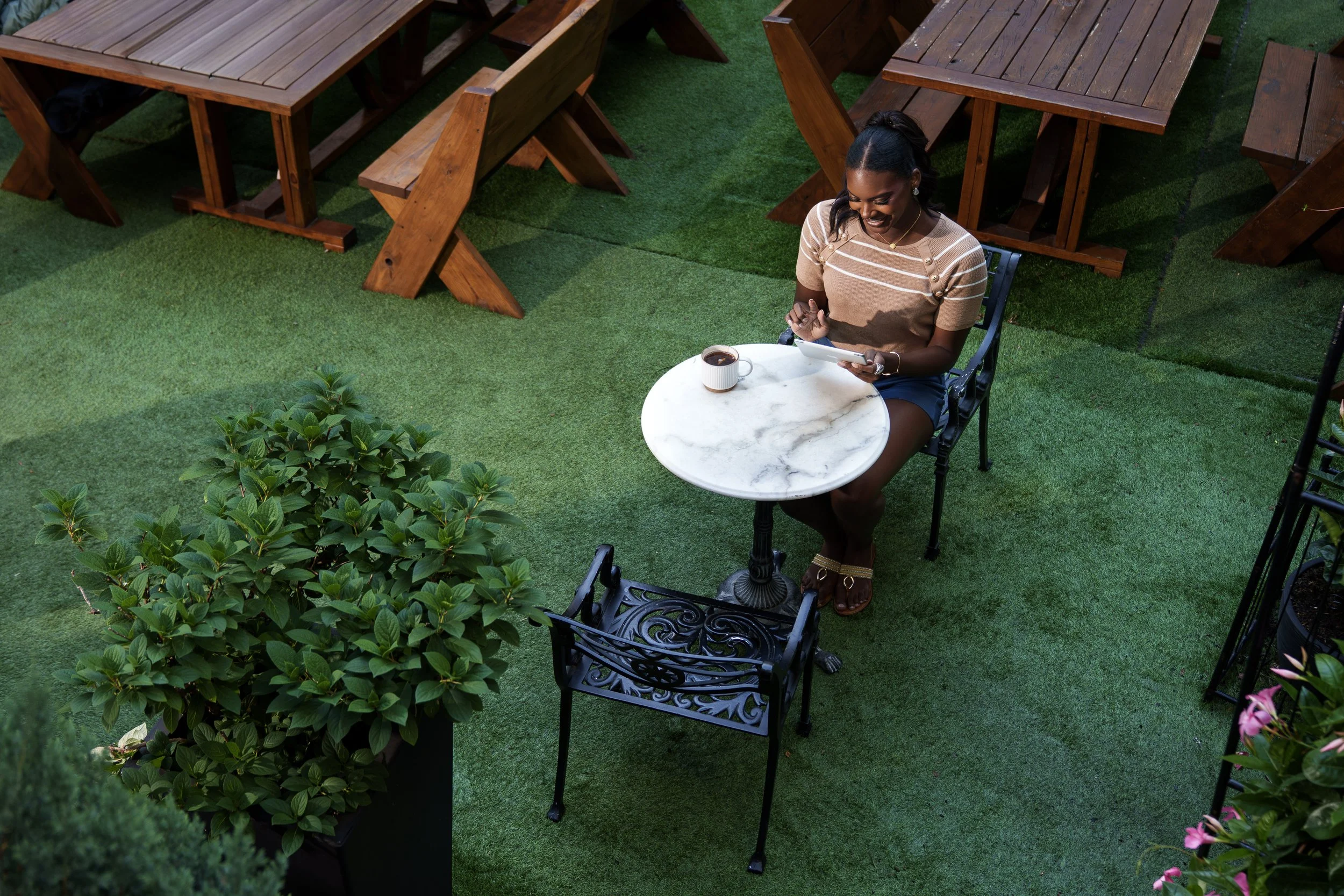 A woman seated at a small round table outdoors, reading on a smartphone with a coffee mug nearby, surrounded by wooden tables and benches on a grassy patio.