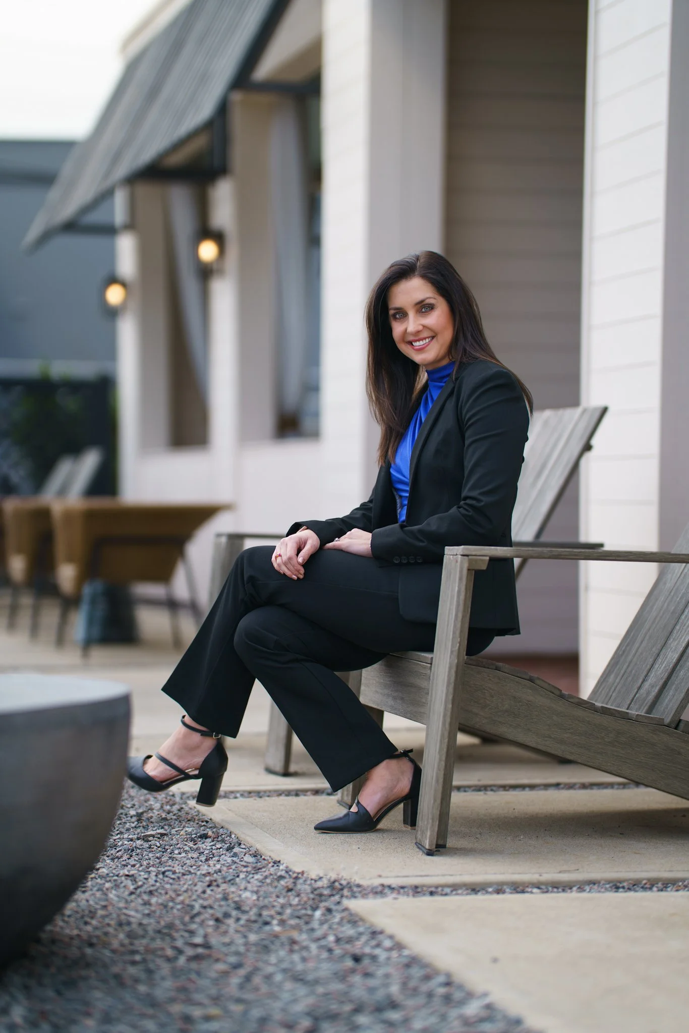 A woman sitting on a wooden lounge chair outdoors, smiling at the camera, wearing a black suit and a blue top with high heels.