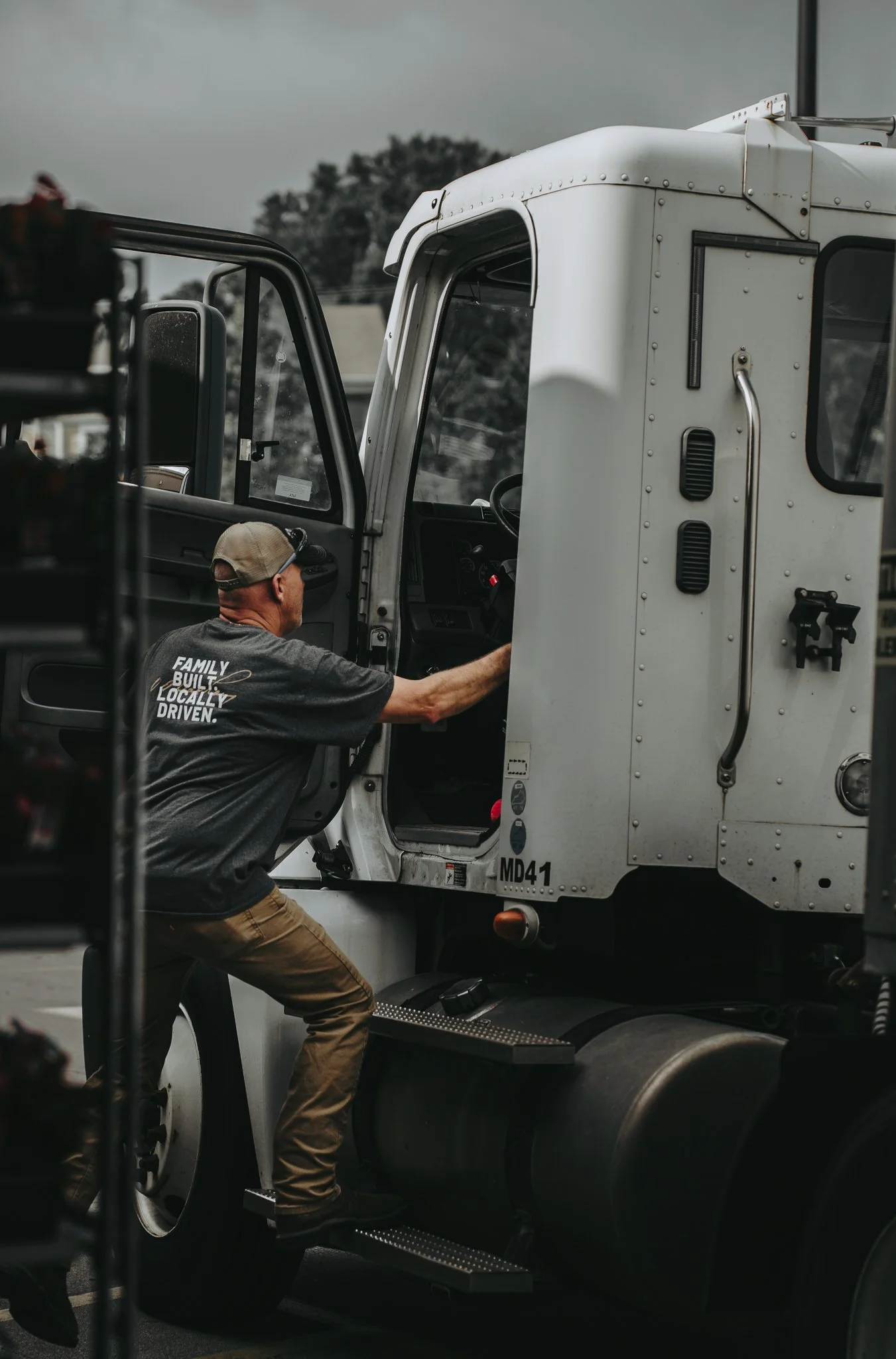 A man wearing a gray T-shirt and tan pants is standing on the edge of a truck, reaching into the driver’s seat.