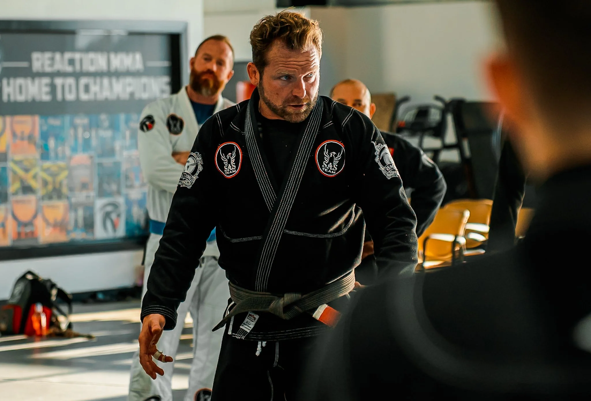 A man wearing a black martial arts gi with patches, a gray belt, and holding a cigar, appears to be instructing or talking to someone outside the frame. Several other men in martial arts gis are in the background, with a chessboard and some sports eq