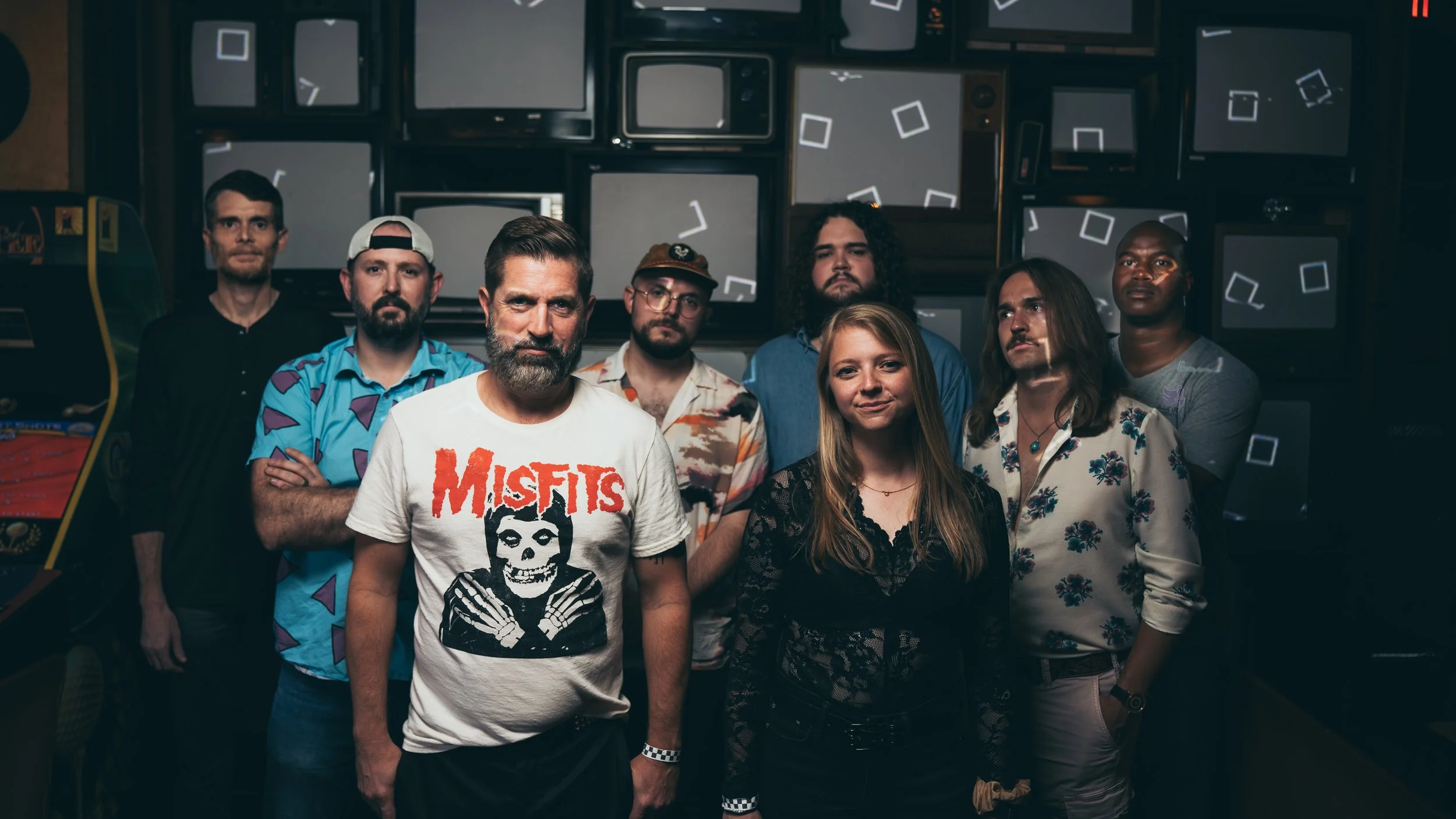 A group of eight people standing in front of a wall of vintage television screens with static and geometric patterns. The group includes six men and two women, with casual and retro clothing styles.