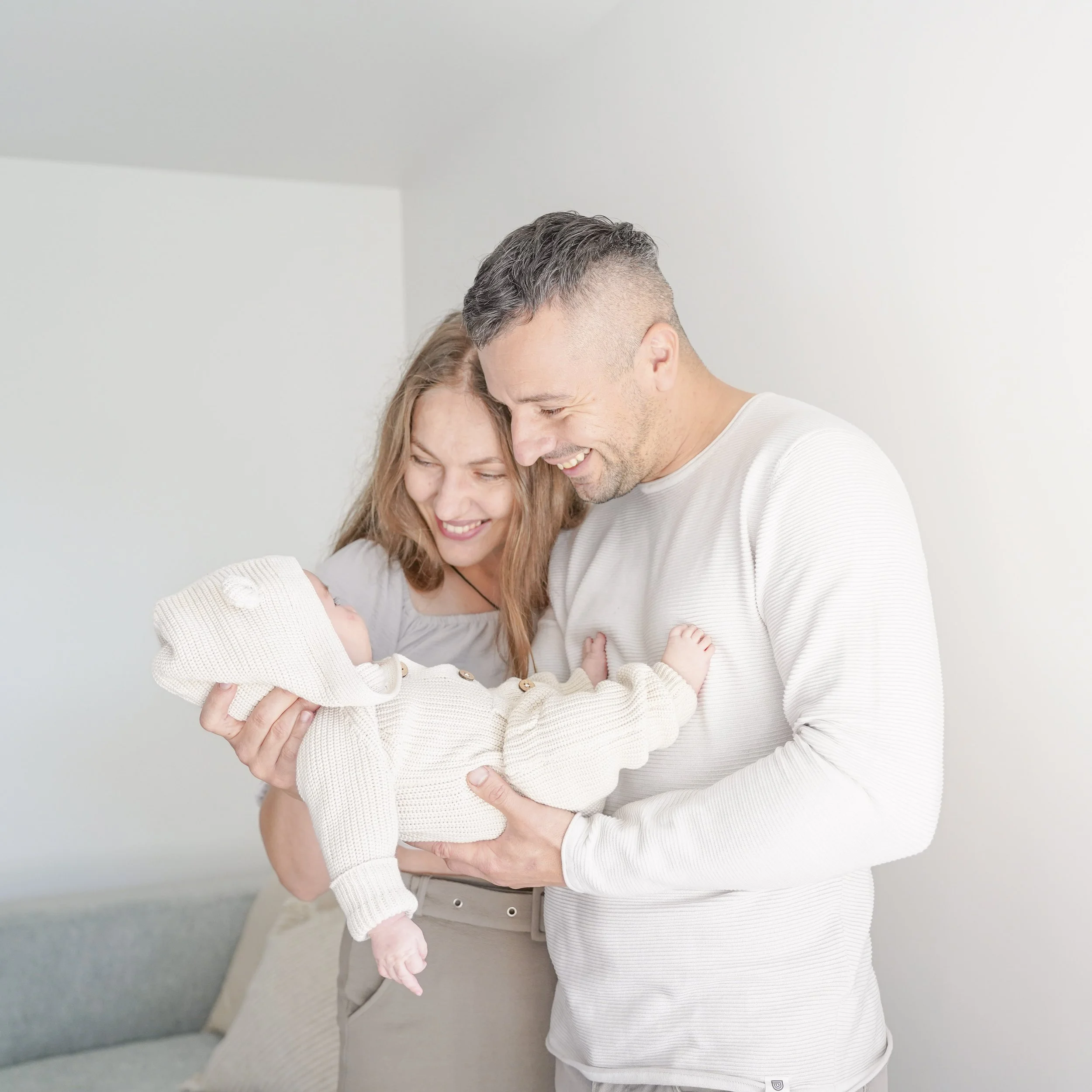 A family of three: a man, a woman, and a baby, all smiling and hugging each other in a bright room.