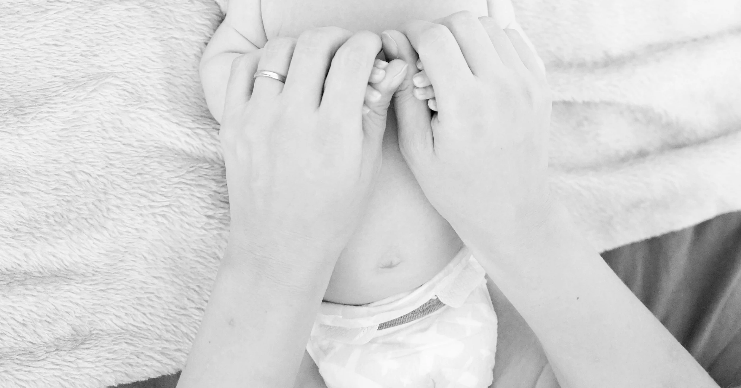 A close-up of a baby holding an adult's finger, with an adult hand supporting the baby's hand, in black and white.