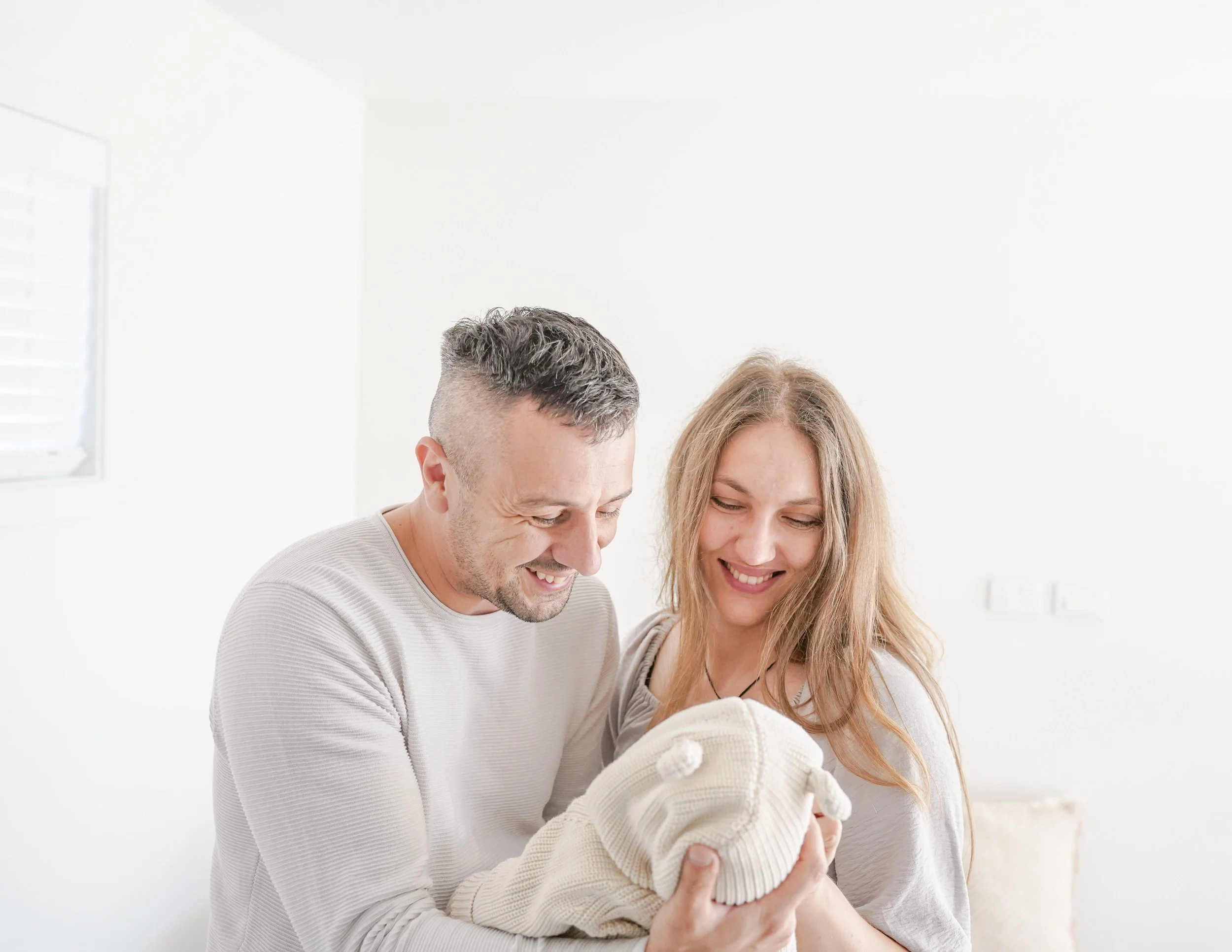 A smiling man and woman looking at a baby in a cream-colored sweater, in a brightly lit room with white walls.