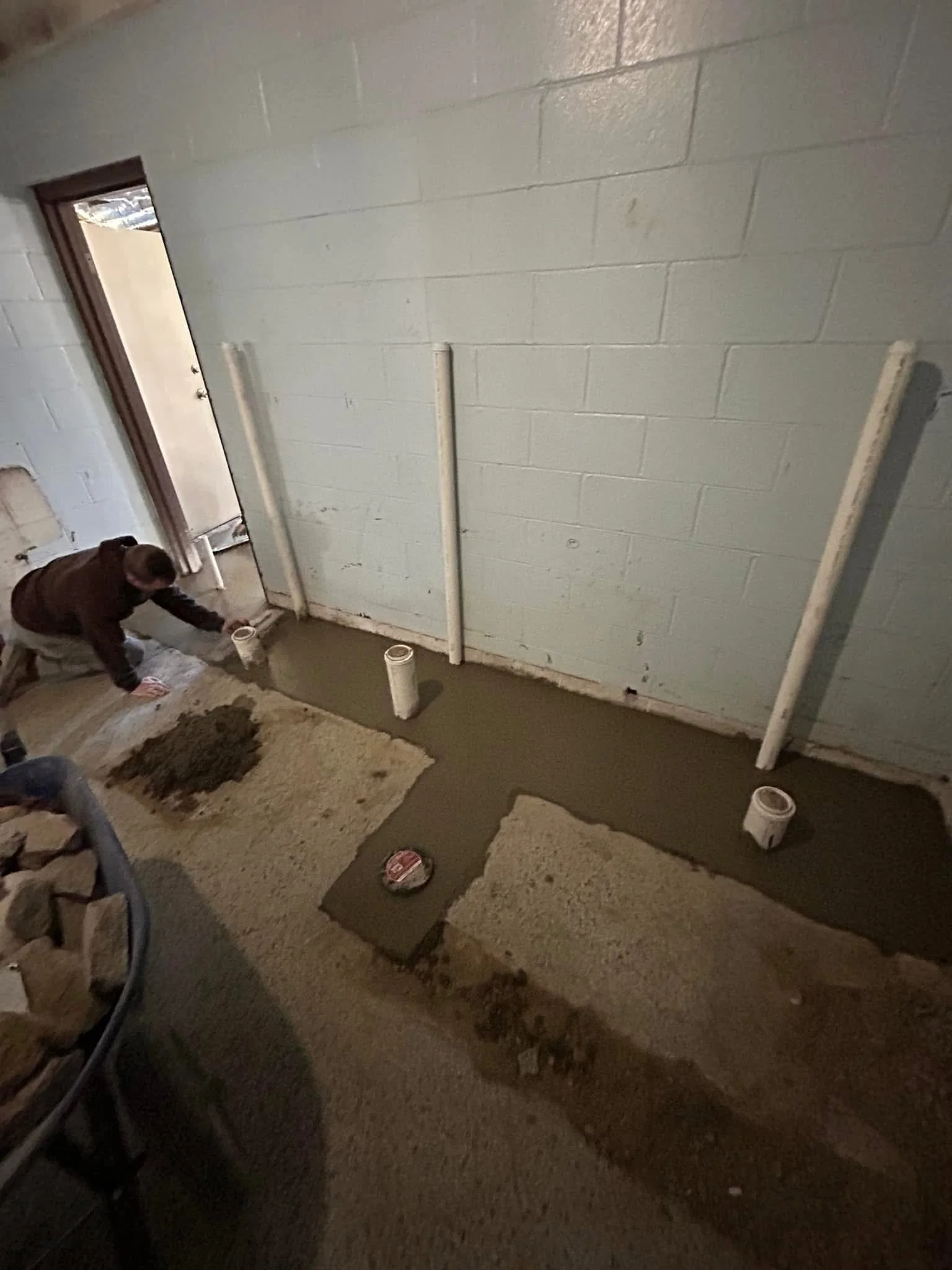 A person working on a home improvement project, laying down a concrete floor inside a room with cinder block walls. Four white pipes protrude vertically from the concrete, and a bucket of cement is on the floor. A doorway leads outside.
