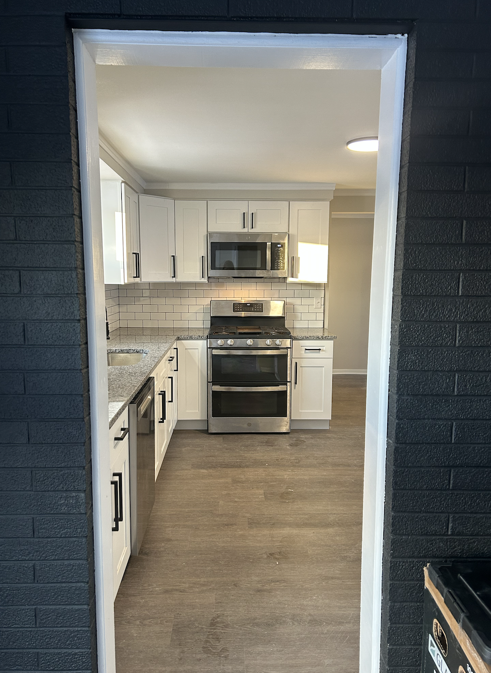 View of a modern kitchen with white cabinets, a stainless steel stove with oven, microwave, granite countertops, and a tiled backsplash, seen through a doorway with black exterior walls.