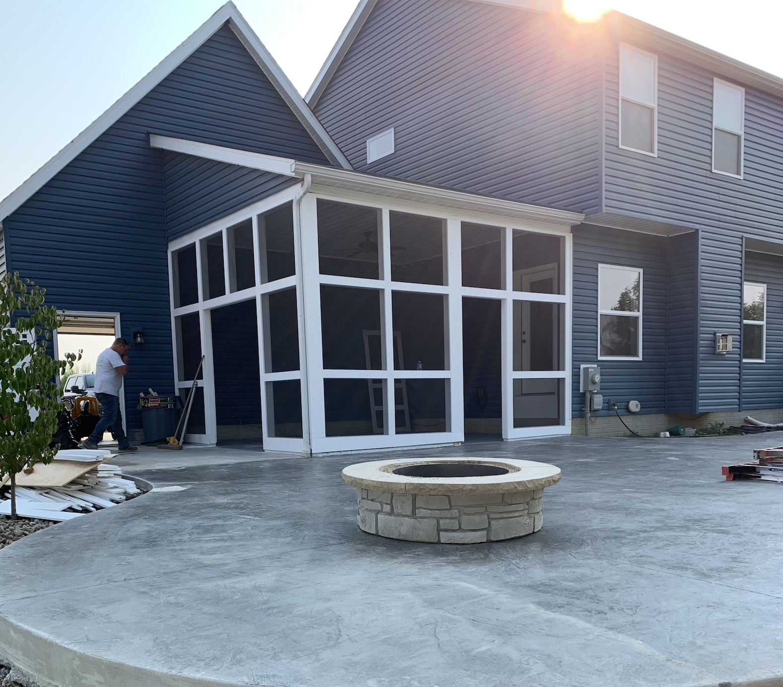 A two-story blue house with a white screened-in porch and a concrete patio. There is a man working on the left side near construction materials, and a stone fire pit in the center of the patio. The sun is shining brightly.