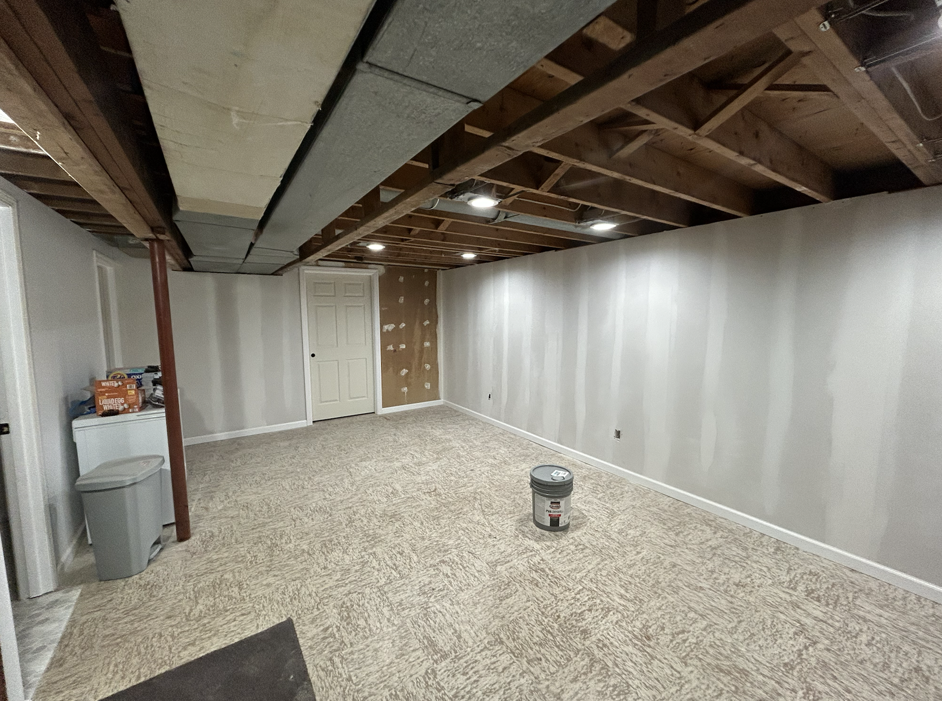 Undeveloped basement room with white painted walls, exposed wooden ceiling beams, and a beige carpeted floor; construction materials and a laundry basket are visible.
