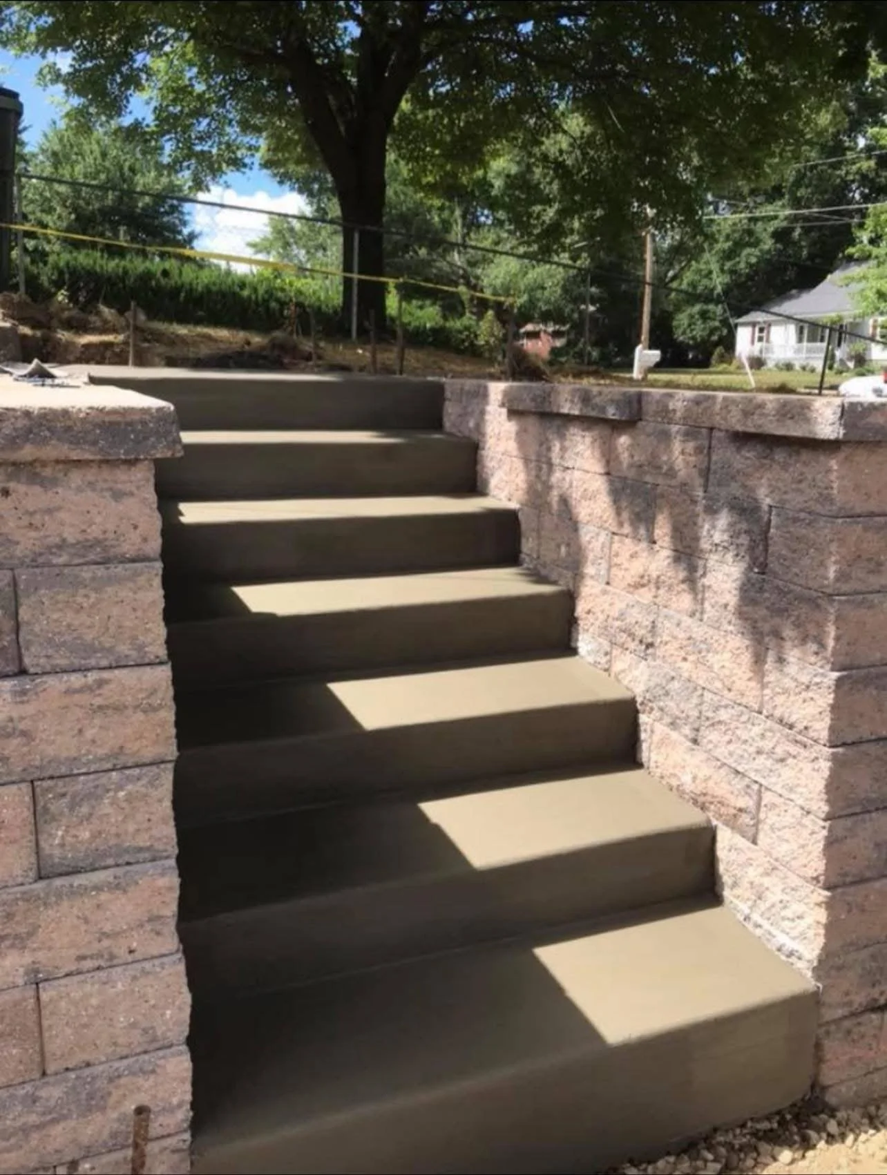 Construction of concrete stairs between two stone walls outdoors, with trees and houses in the background.