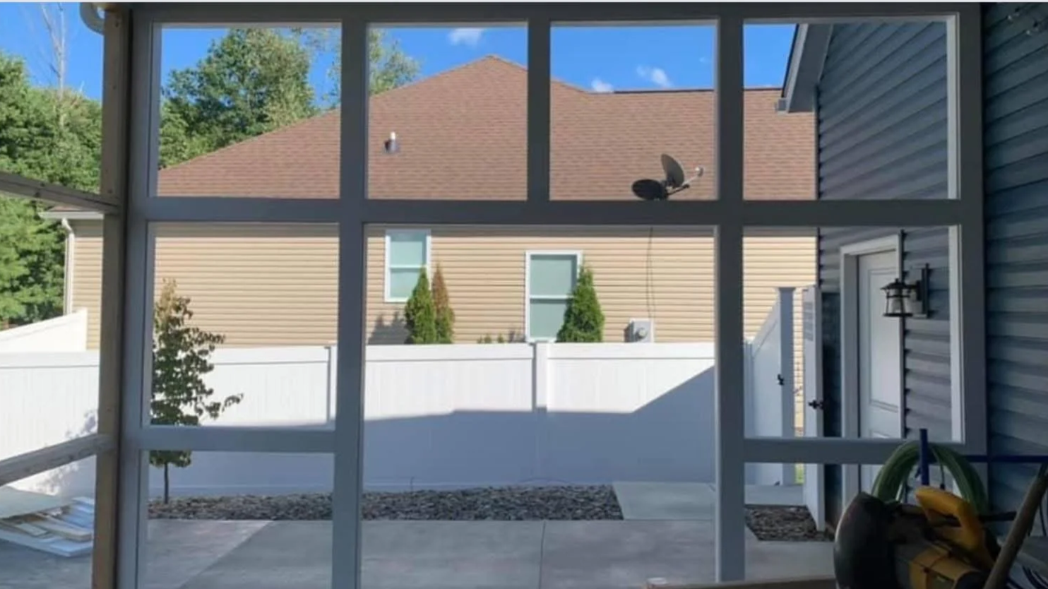 View through a screened porch looking out onto a backyard with a white fence, two small trees, and a house with beige siding and a brown roof.