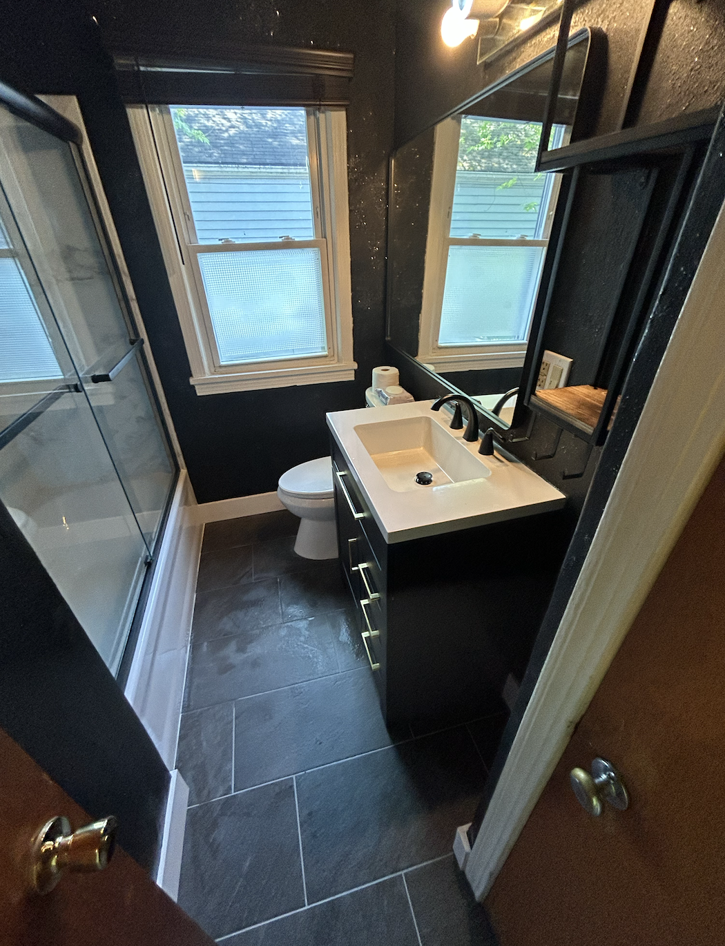 Small modern bathroom with dark walls, large window, shower with glass doors, white sink with black fixtures, mirror, and a toilet.