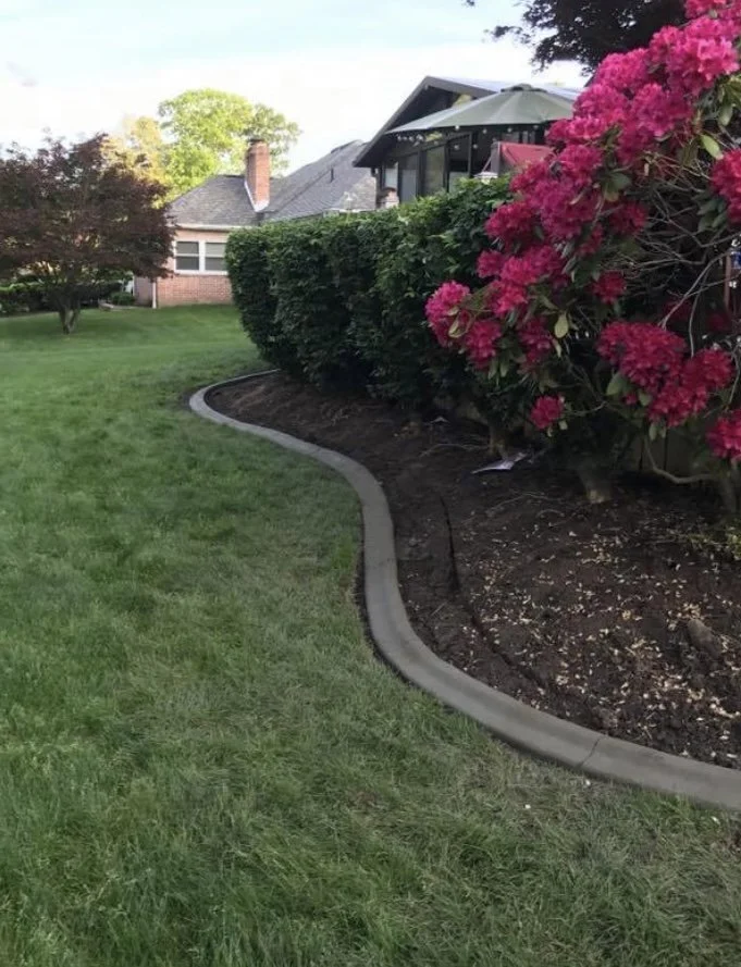 A well-maintained yard with green grass, freshly laid concrete edging, and pink flowering bushes beside a house with a screened porch and patio umbrella.