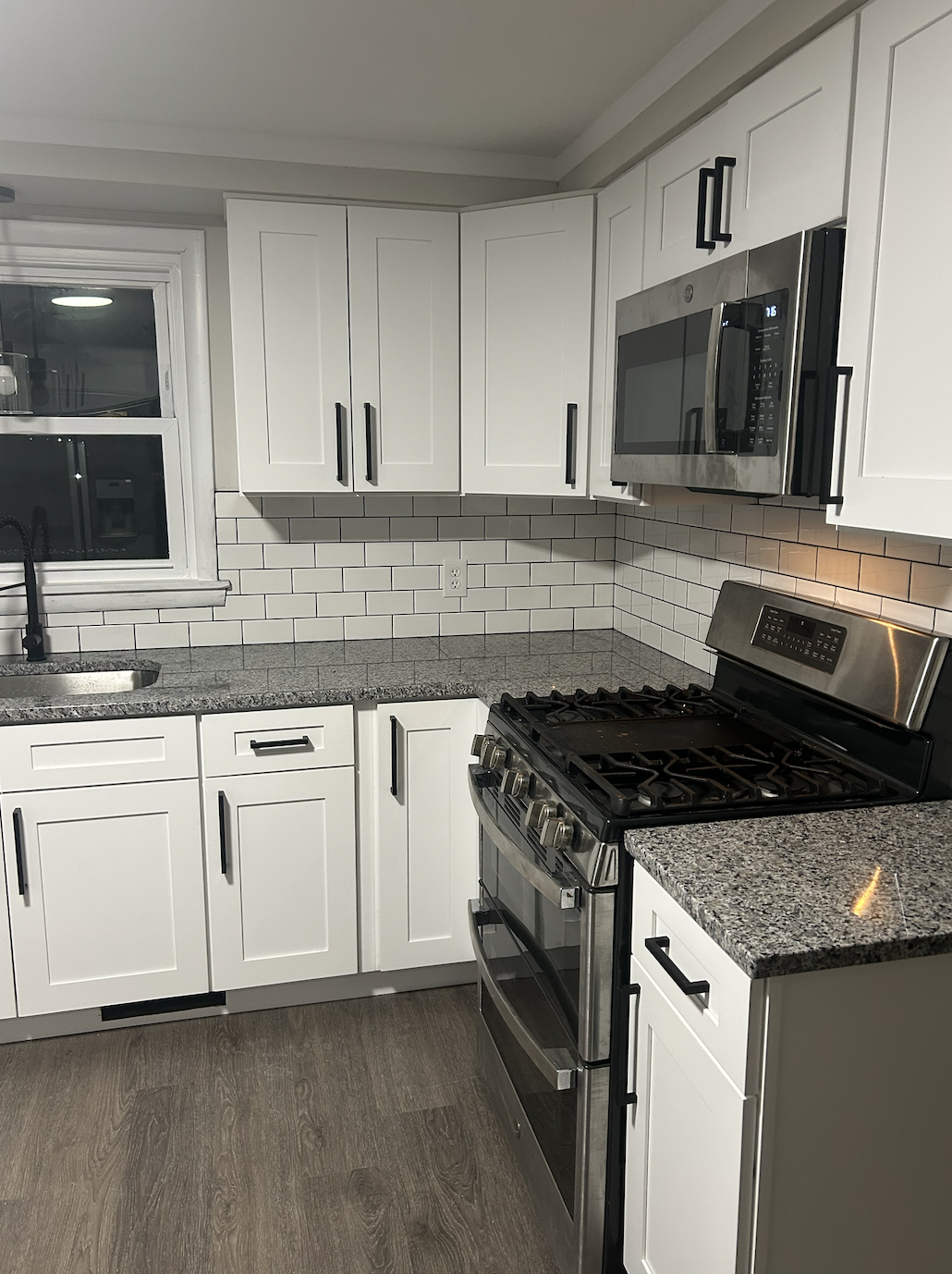 Modern kitchen with white cabinets, granite countertops, a stainless steel microwave above stove, black handles on cabinets, white subway tile backsplash, window above sink, and a dark wood floor.