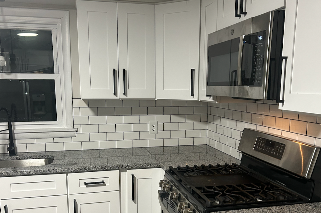 Kitchen with white cabinets, gray granite countertop, subway tile backsplash, stainless steel microwave, stove, and a window above the sink.