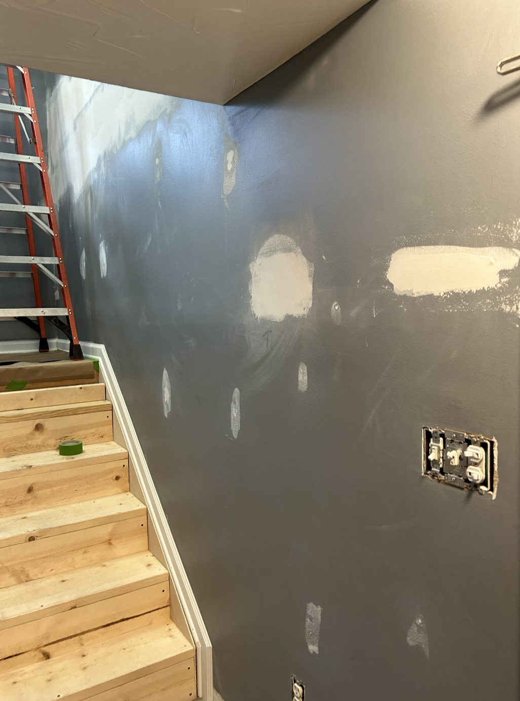 An interior wall undergoing renovation, with patches of drywall and paint, electrical outlets, and an unfinished staircase with wooden steps and a red ladder nearby.